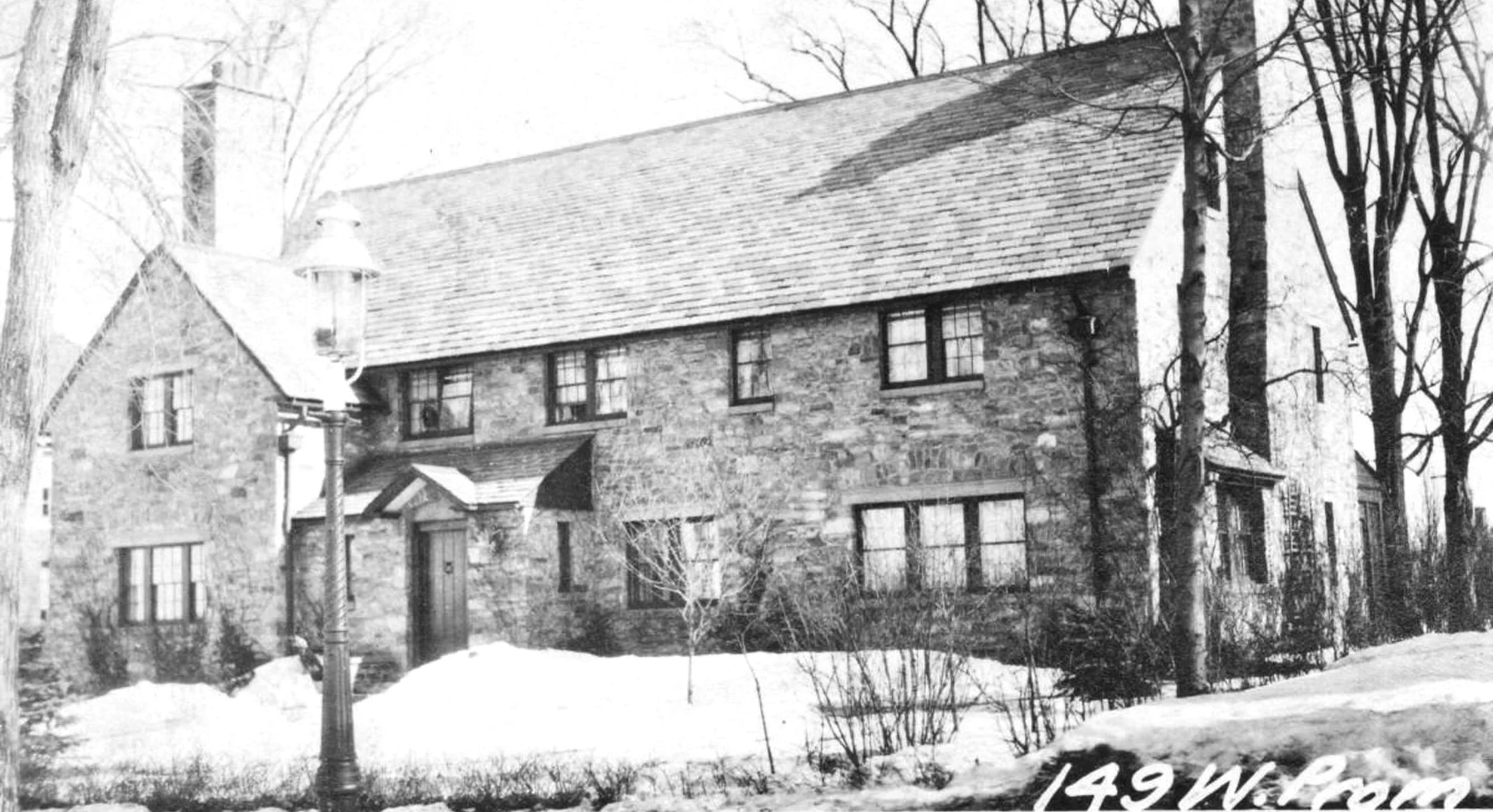 Black and white photo of a two-story stone house with a steep roof, surrounded by bare trees and snow on the ground.