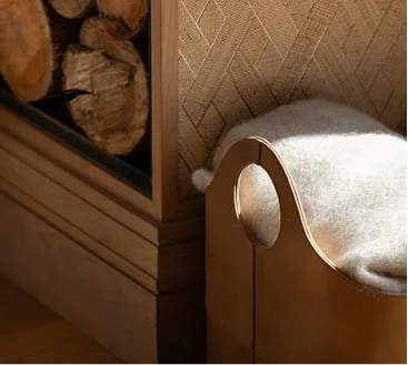 A corner of a wooden piece of furniture with a circular cutout and plush white fabric, next to a wall with a textured pattern and a stack of logs.