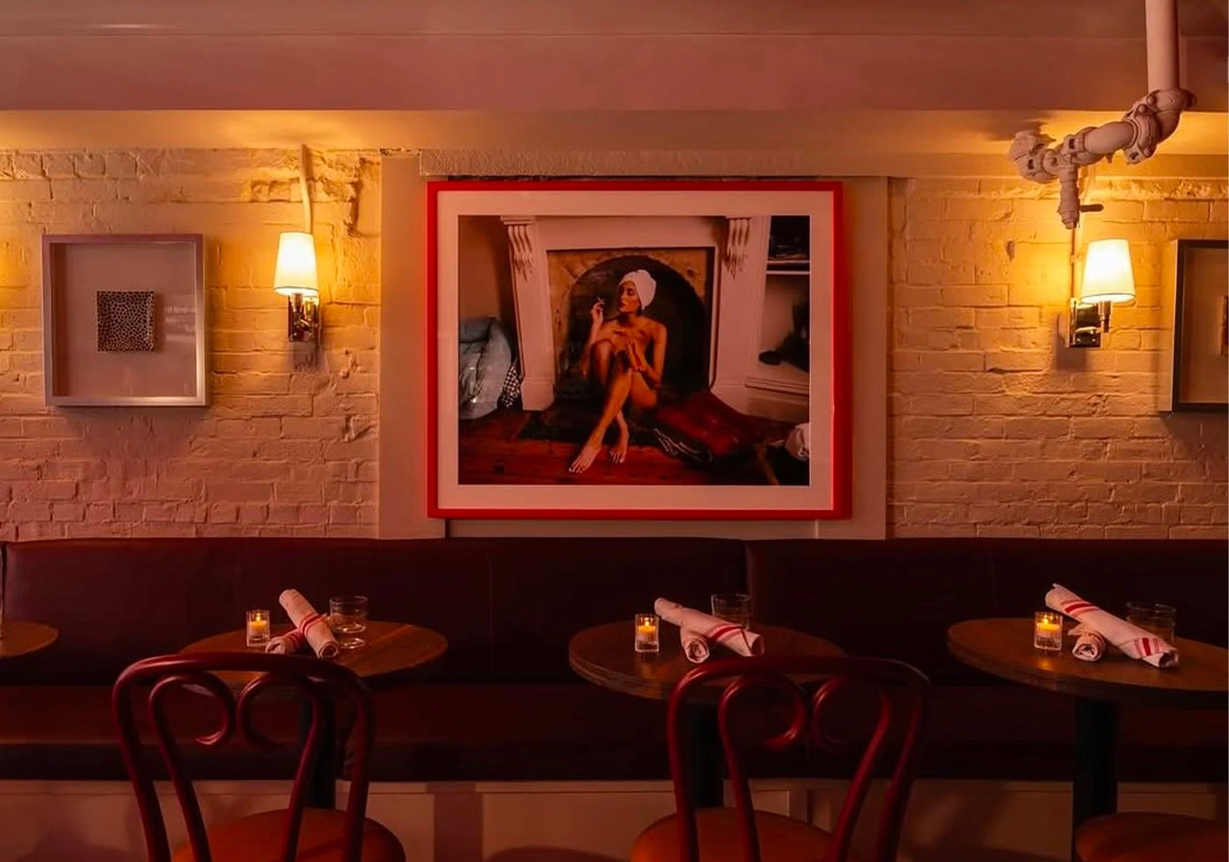 Interior of a restaurant with brick walls, wall lamps, and a framed artwork of a woman smoking a cigarette, with tables set for dining including candles and rolled napkins.