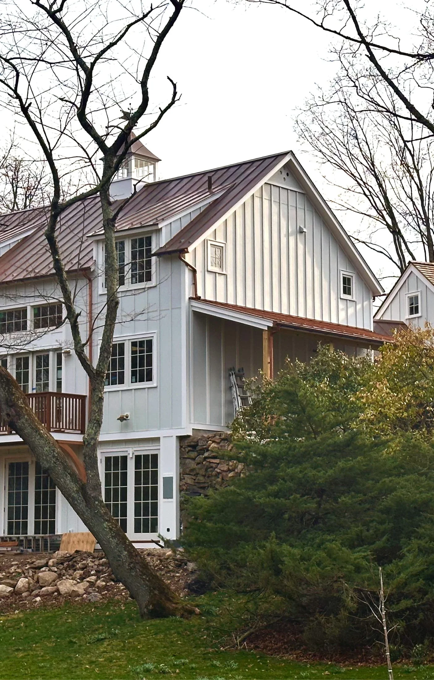 A two-story house with white wooden siding and a metal roof under an overcast sky, surrounded by leafless trees and green bushes, with rocks at the base of the house.