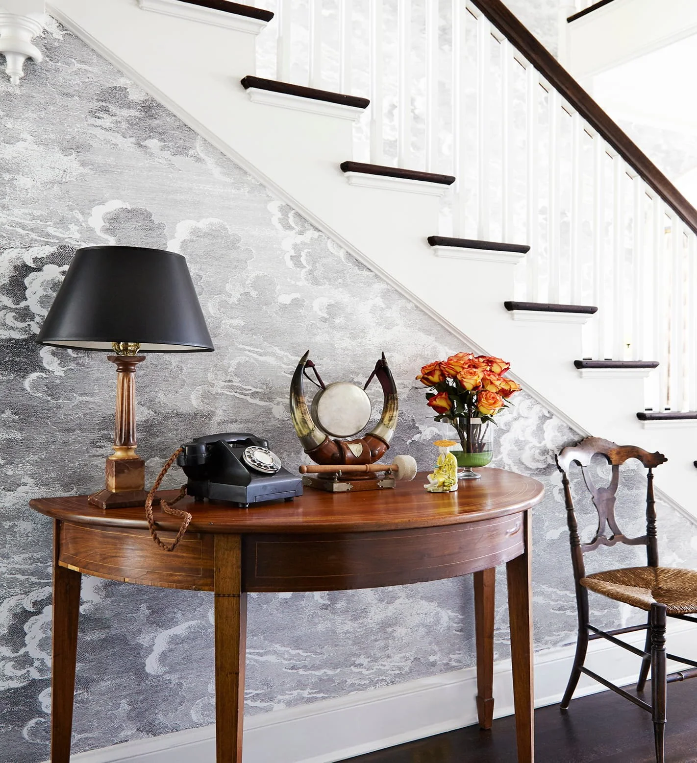 A vintage black rotary phone and a decorative drum mirror on a wooden desk, with a black lampshade, a glass vase of orange roses, and a small yellow Buddha statue. A carved wooden chair with a woven seat is beside the desk, under a staircase with white spindles and dark wooden steps, against a textured grey and white wallpapered wall.