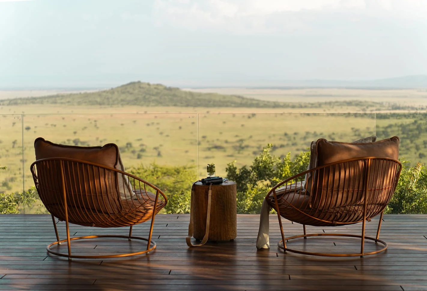 Two modern outdoor chairs with leather cushions and metal frames sit on a wooden deck, overlooking a vast, open savannah landscape with a distant hill and scattered trees under a partly cloudy sky.