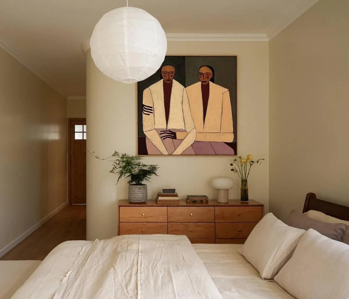 A bedroom with a bed covered in white bedding and pillows, a wooden dresser with decorative items including potted plants, a lamp, and books, a large abstract painting of two seated individuals hanging above the dresser, and a hanging paper lantern light fixture. The room has beige walls and a wooden door in the background.