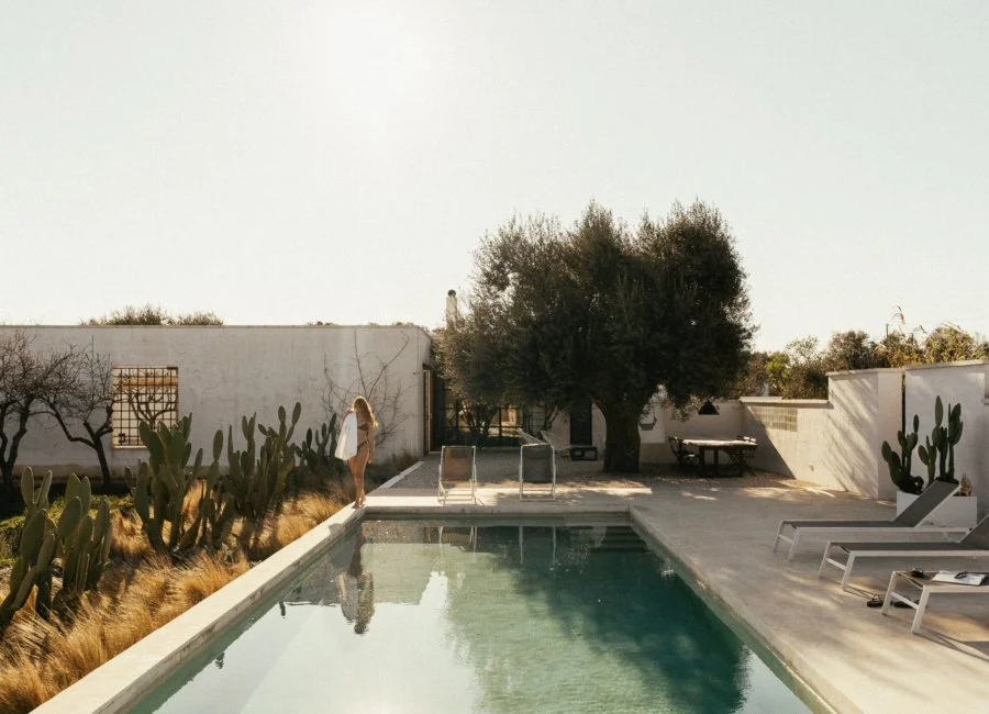 A peaceful backyard scene with a swimming pool, white lounge chairs, cacti, a large tree, and a woman standing near the poolside.