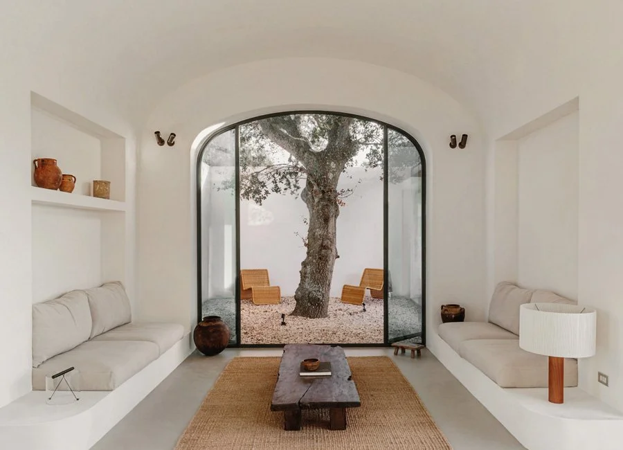 Living room with white walls, built-in couches, a wooden coffee table, and a large glass door revealing an outdoor space with a tree and two chairs.