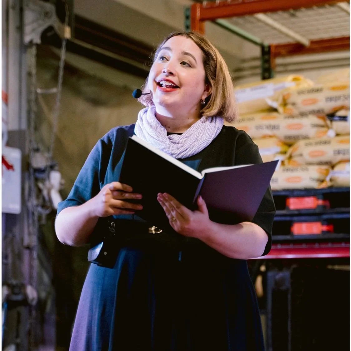 A woman with shoulder-length brown hair wearing a black dress, a light purple scarf, and a headset microphone, speaking or presenting in an industrial or warehouse setting with sacks of supplies stacked behind her.
