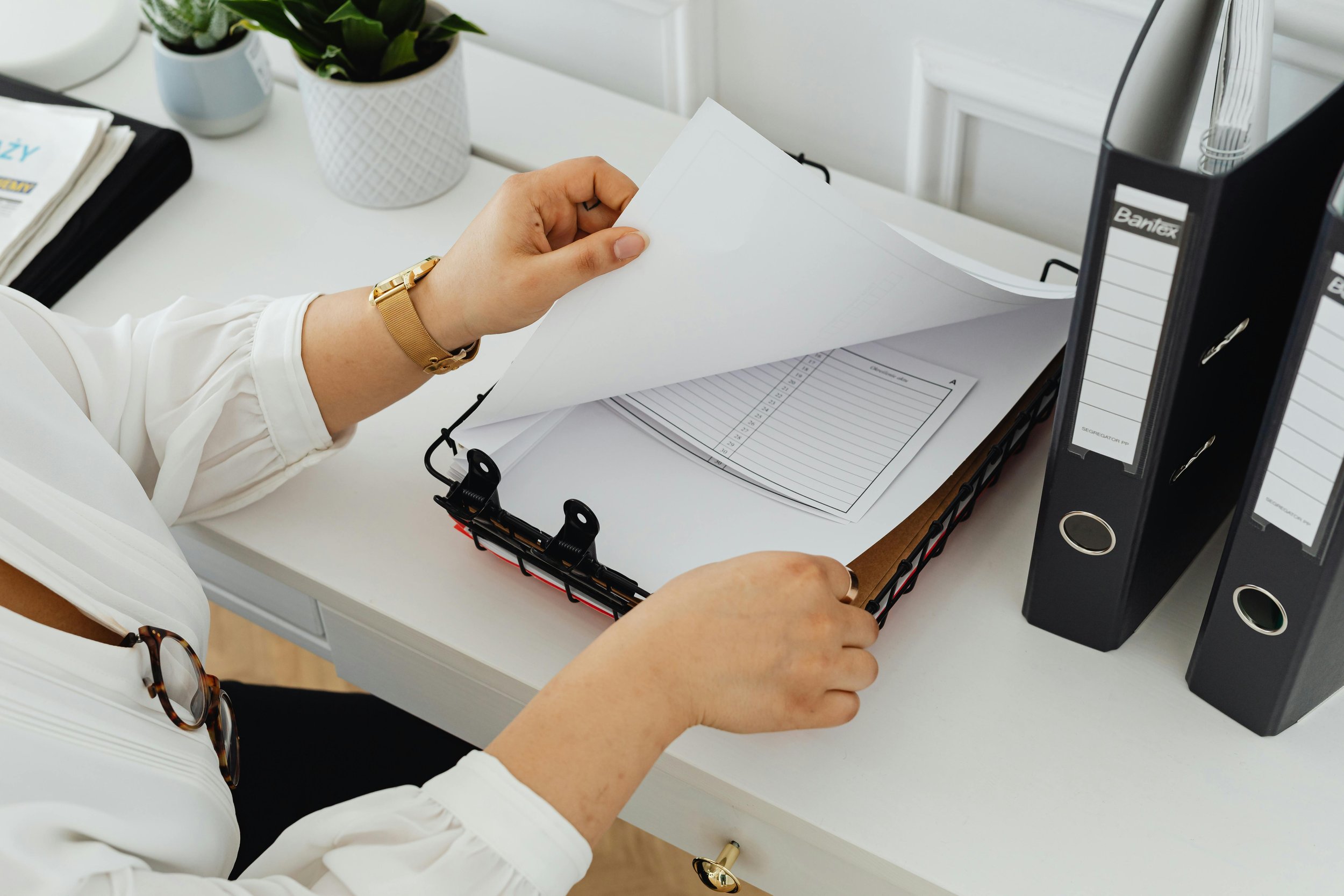 A person organizing papers in a black wire tray on a white desk, with binding files and potted plants nearby.