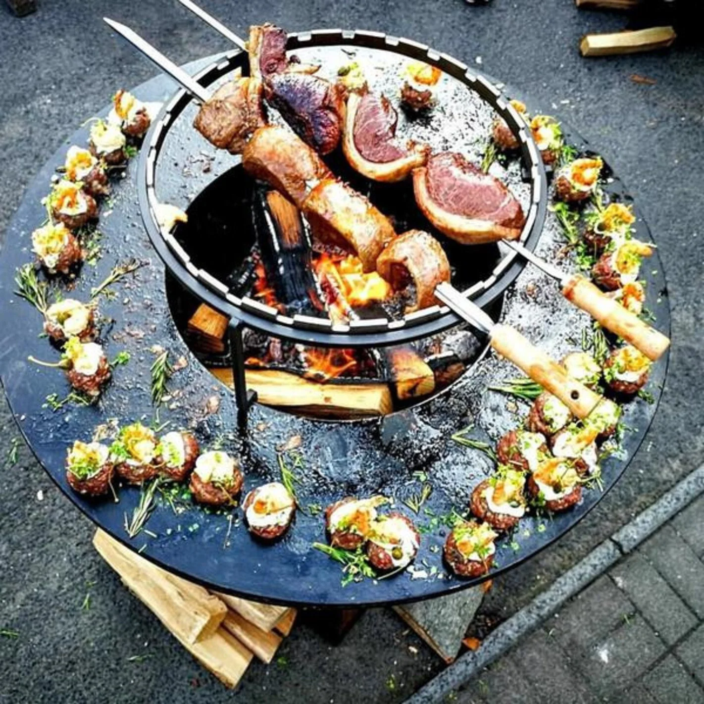 Une table ronde avec des morceaux de viande grillée, entourée de petits amuse-bouches garnis et décorés avec des herbes, le tout sur un grill au-dessus de flammes.