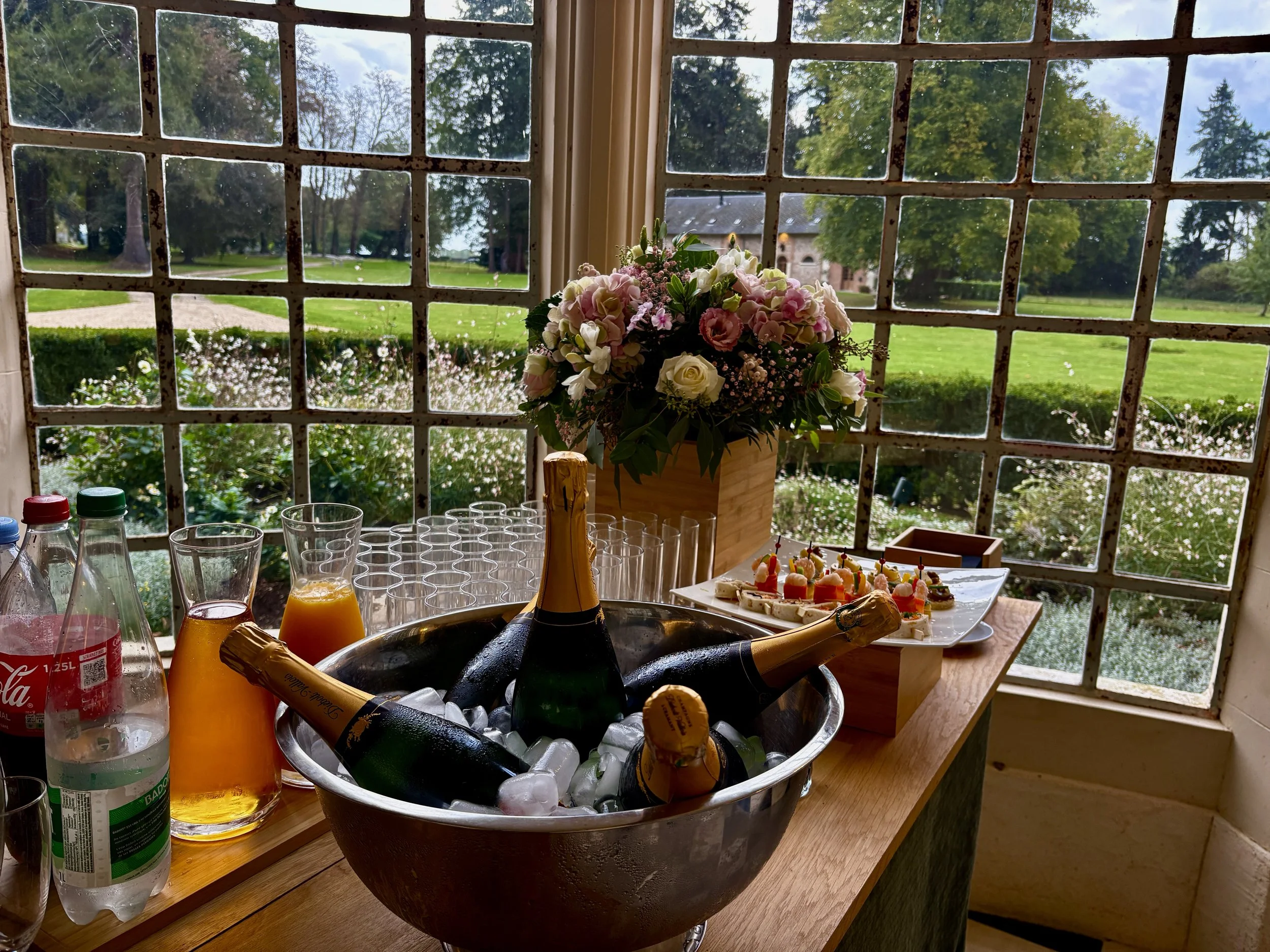 Table avec une bouteille de champagne, des verres, une bouteille de champagne dans un seau à glace, des bouteilles de boissons et une assiette de petites bouchées, vue à travers une fenêtre avec un paysage de jardin et d'arbres.