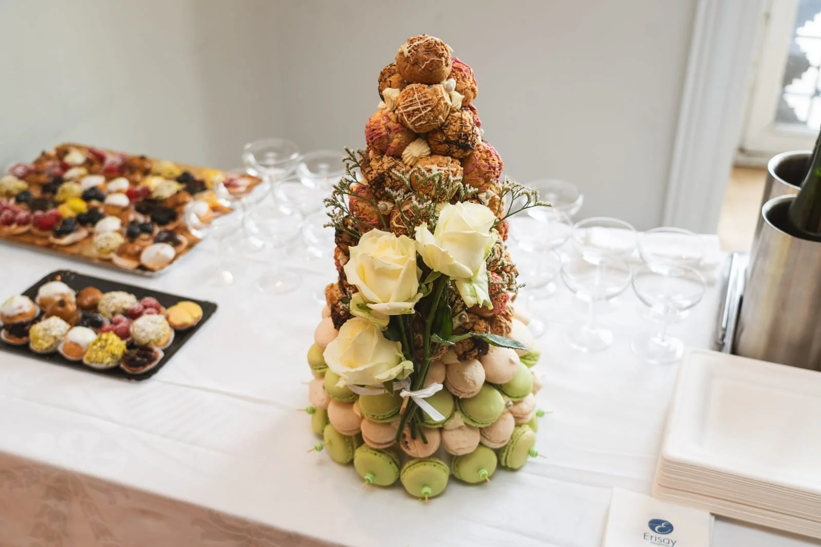 Une pyramide de macarons décorée avec des fleurs blanches, entourée de petits desserts et de flûtes à champagne sur une table, pour une célébration ou un événement.