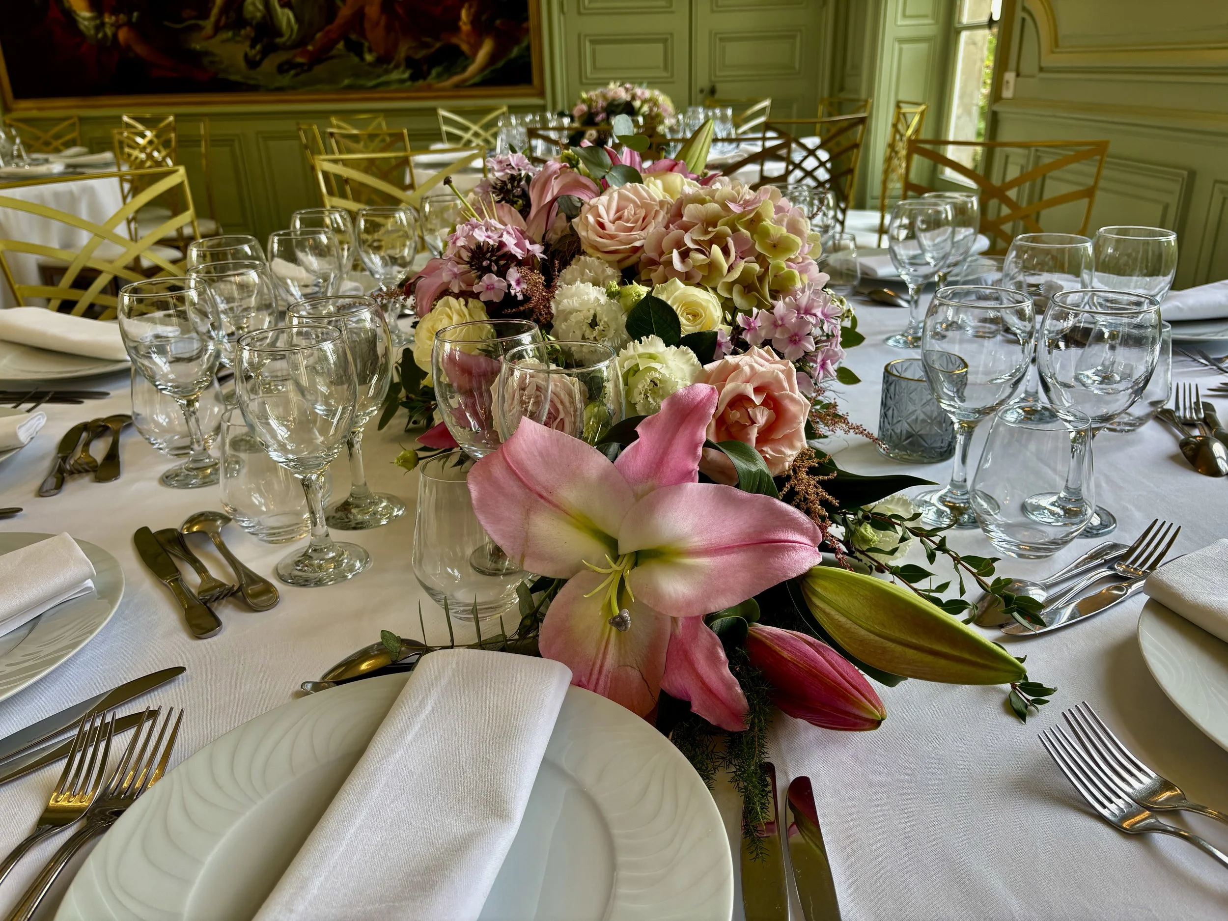 Table de banquet élégamment dressée avec un centre de fleurs, verres à vin, assiettes, fourchettes et couteaux en or, dans une salle décorée de murs verts et d'une peinture.