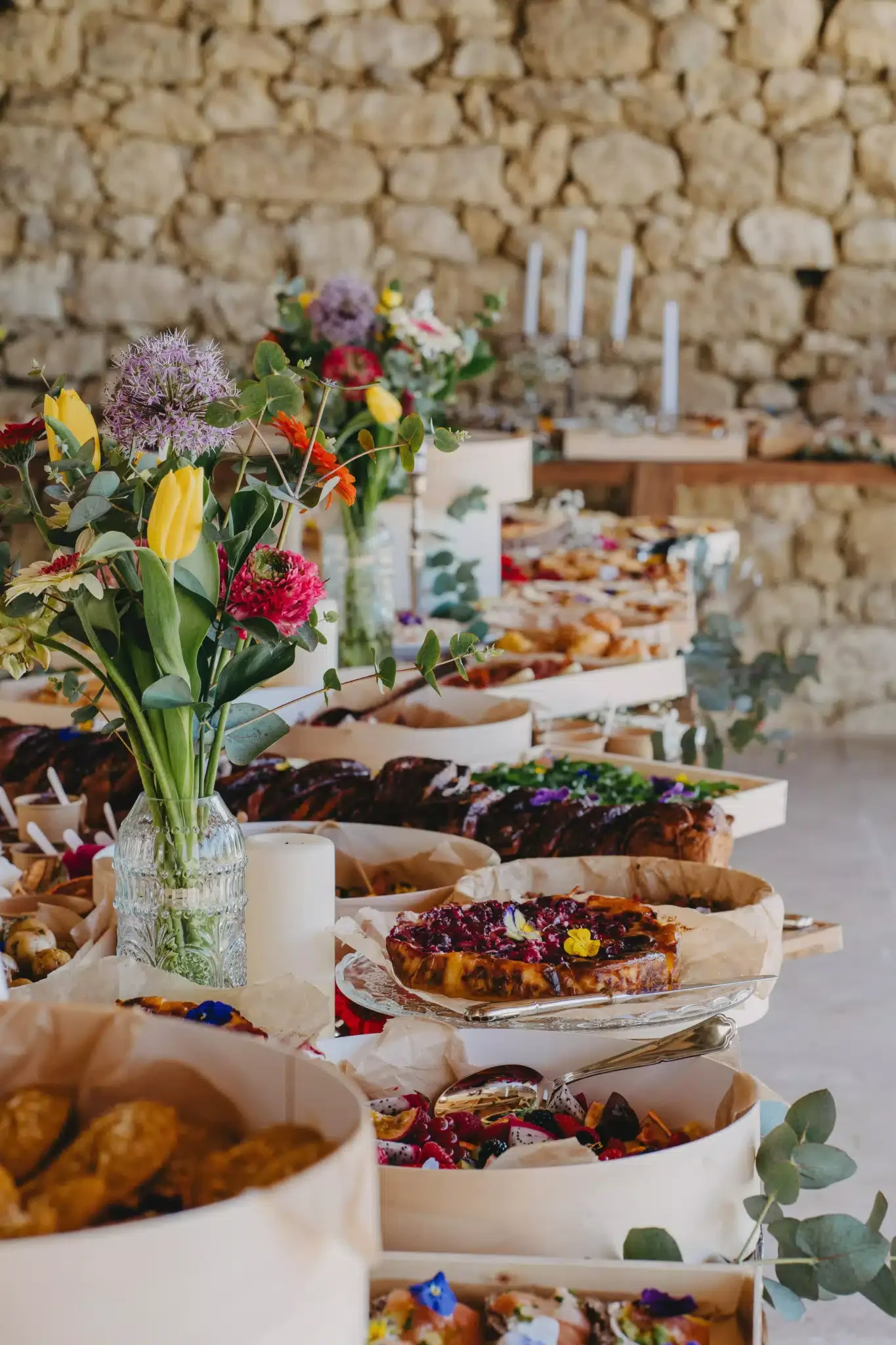 Un arrangement floral dans un vase en verre, avec des fleurs de différentes couleurs, au premier plan, sur une table avec des desserts comme une tarte aux fruits et des petits gâteaux, en arrière-plan une table avec des chandeliers et une pierre