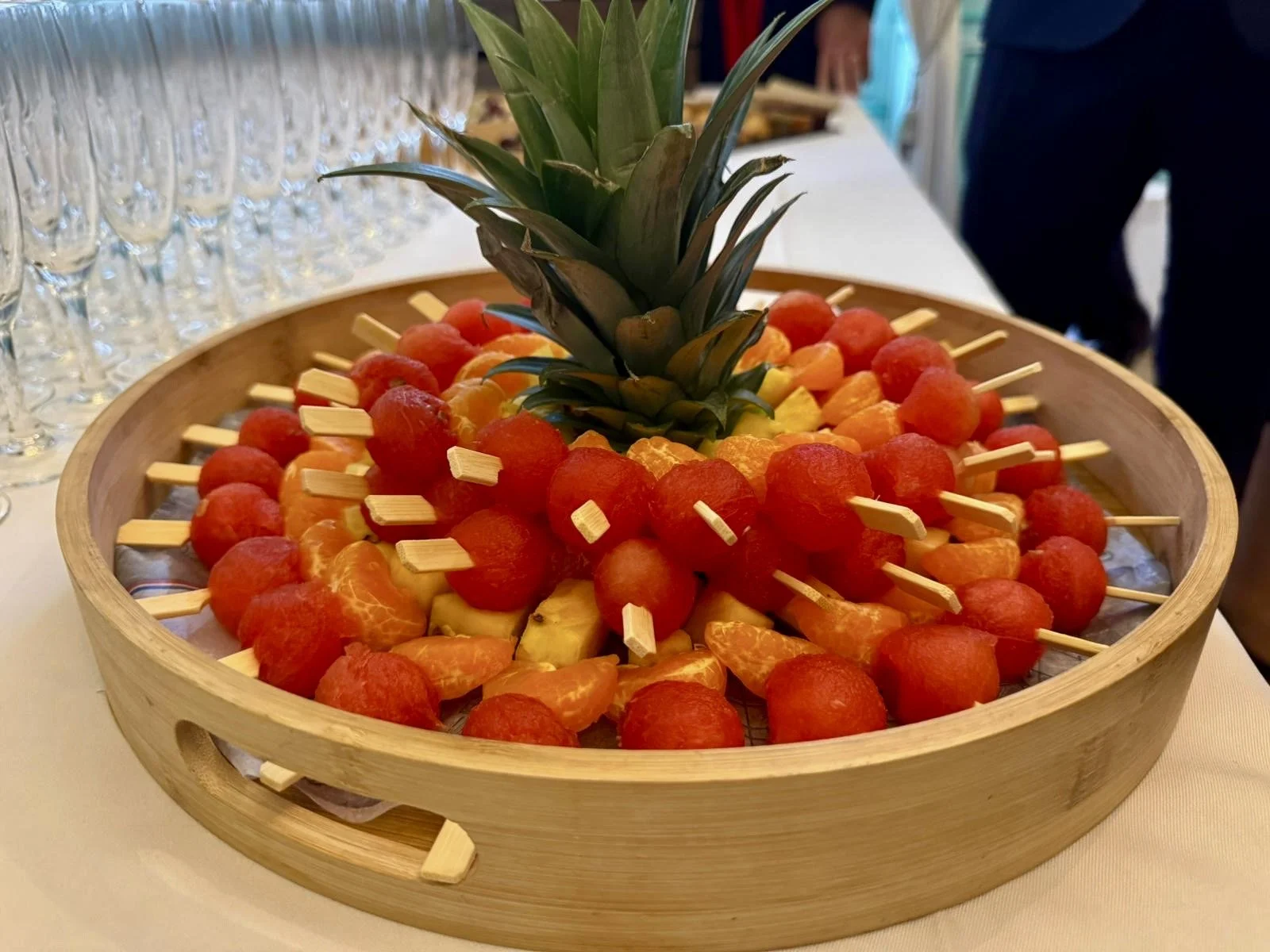 Une grande assiette en bois avec un ananas au centre et des brochettes de morceaux de pastèque, d'ananas, et d'autres fruits.