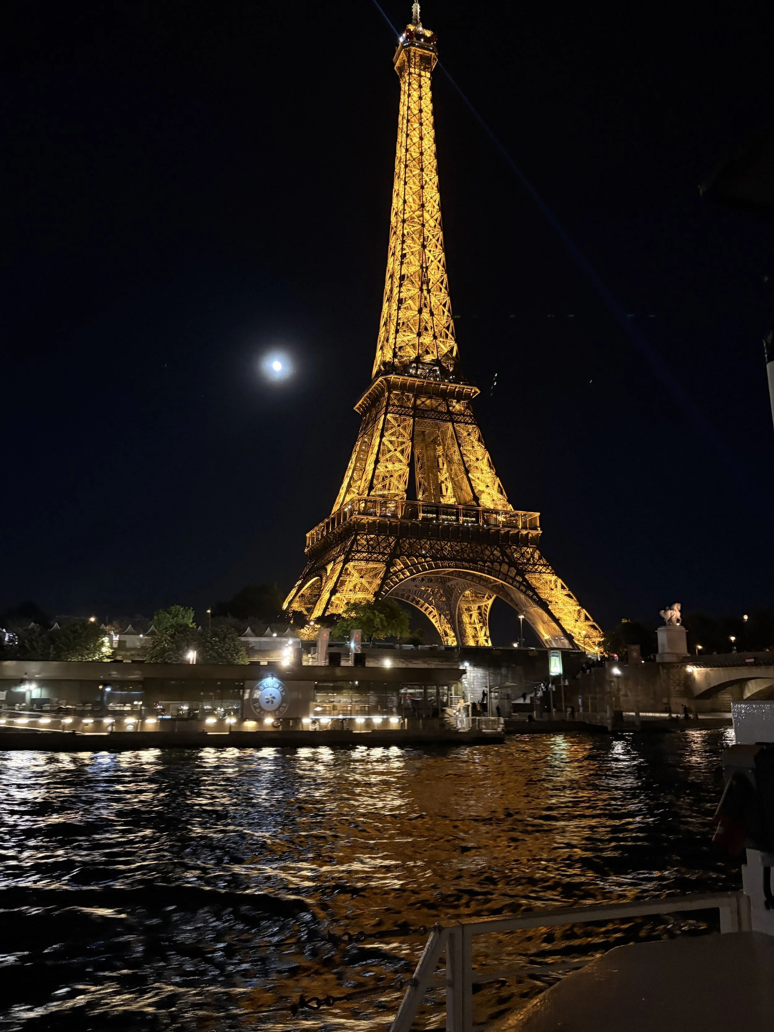 La tour Eiffel illuminée la nuit, vue depuis la Seine, avec la pleine lune dans le ciel et des lumières réfléchies sur l'eau.