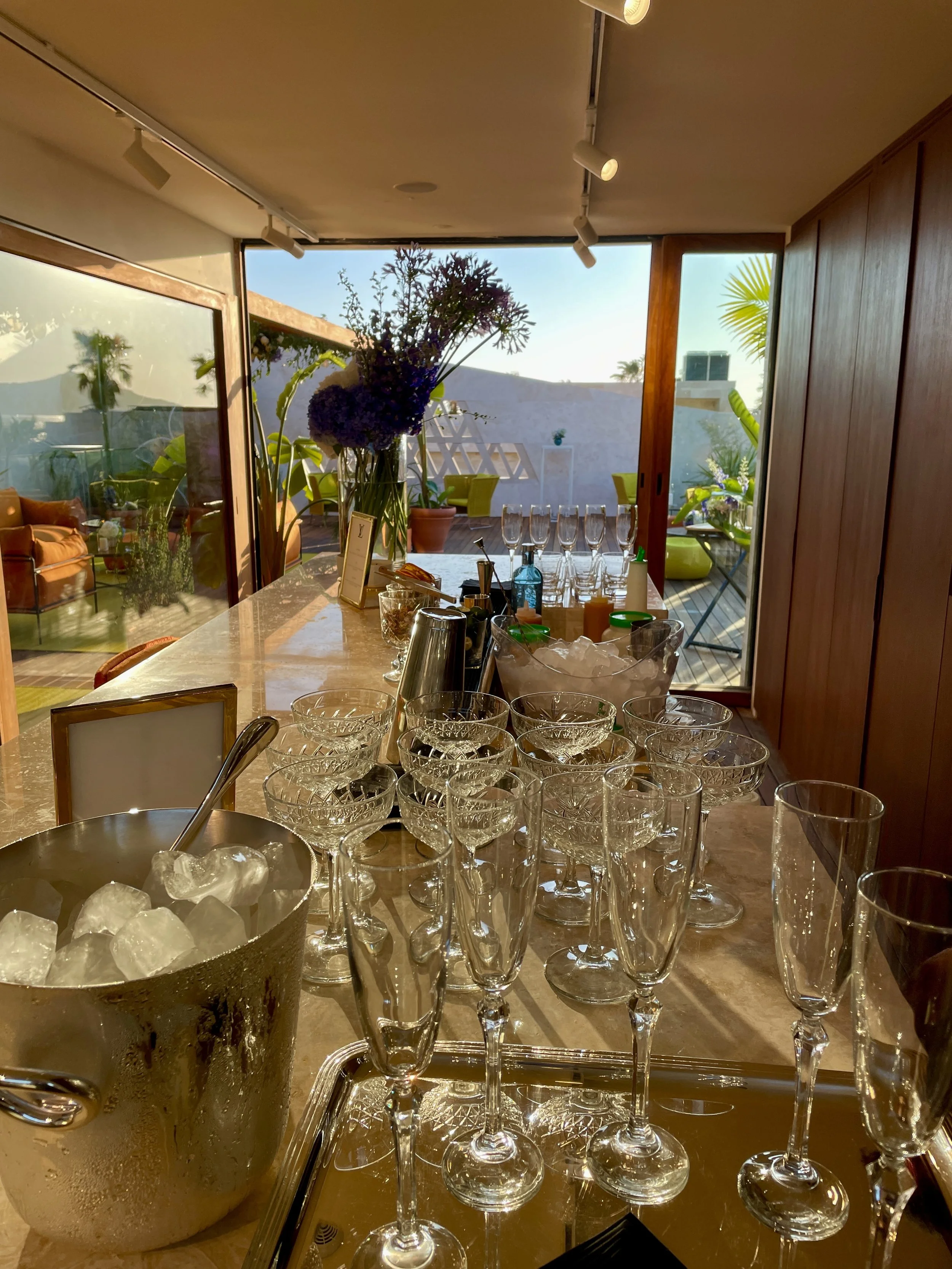 Verre à vin, verres à eau alignés sur une table en marbre, bouquet de fleurs violettes dans un vase, cuisine intérieure avec vue sur un patio extérieur ensoleillé, meubles modernes en bois, plantes dans des pots en dehors.