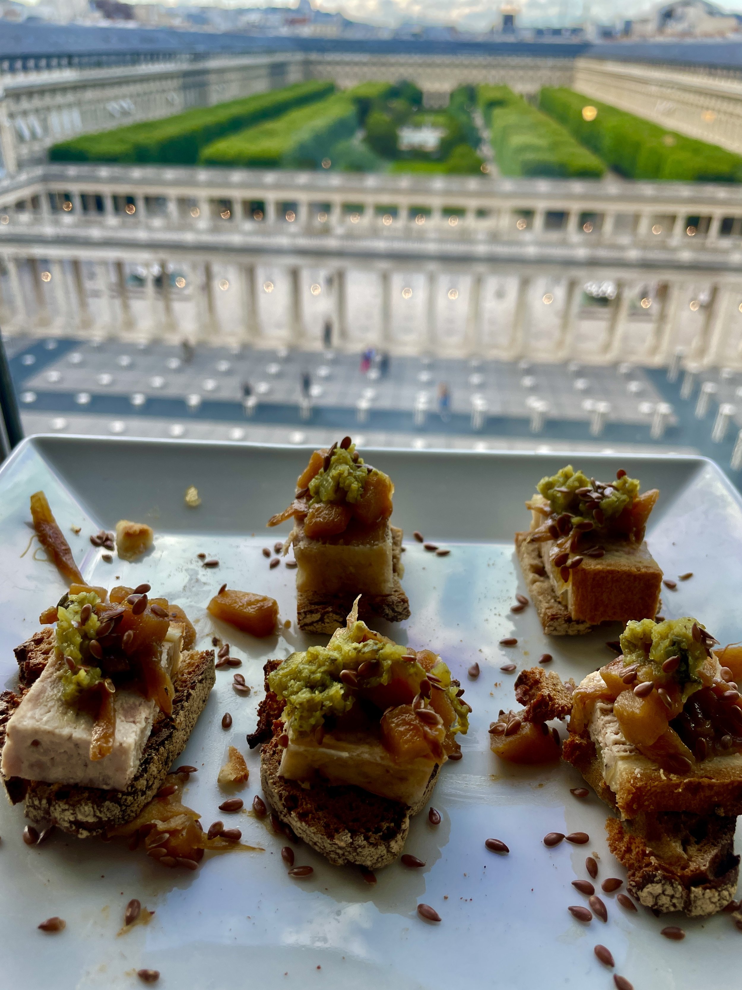 Plate de petits morceaux de pain ou de gâteau garnis de purée d'avocat, de fruits confits et de graines de lin, avec un bâtiment en arrière-plan.