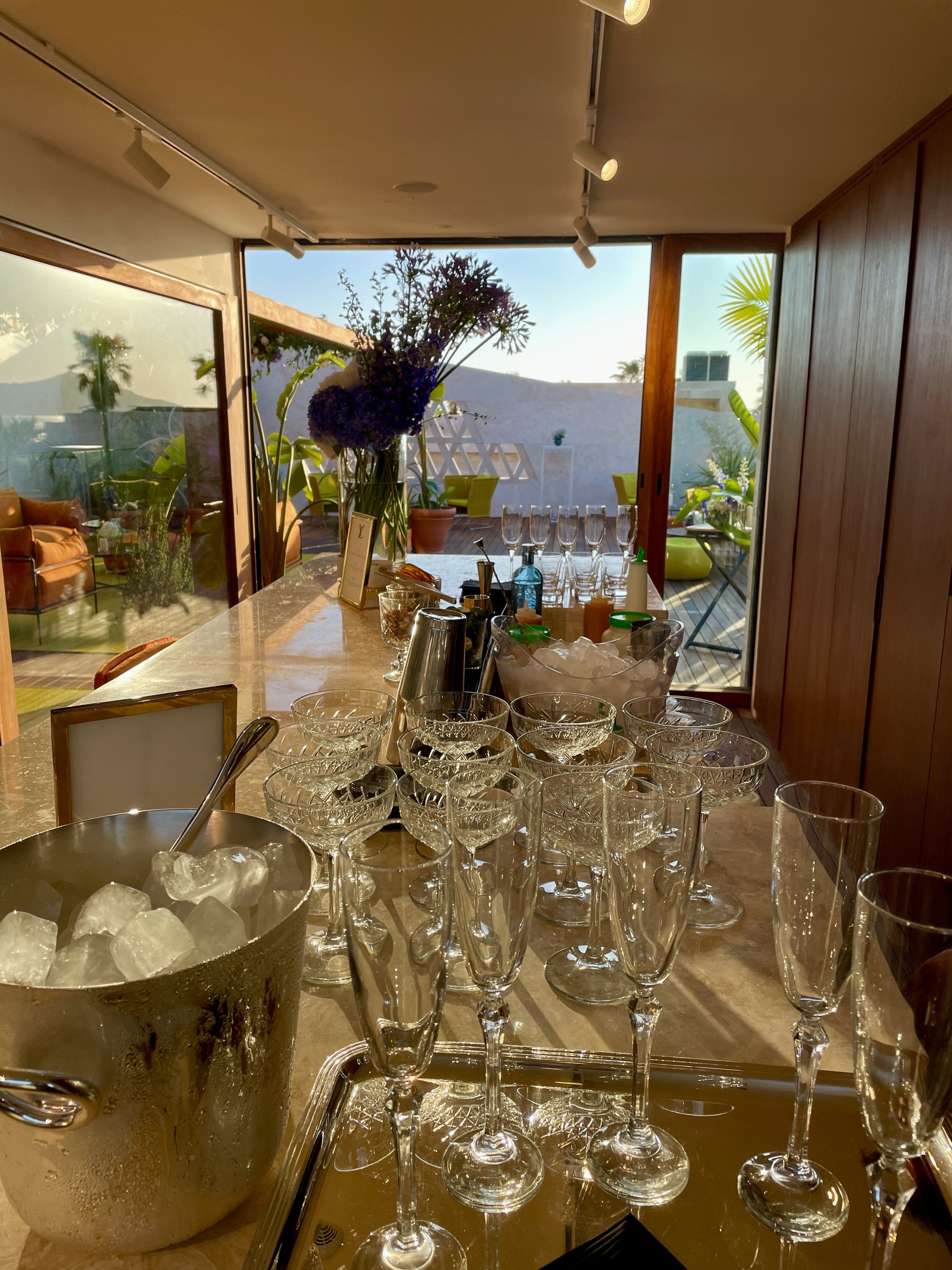 Verres à champagne et un seau à glace avec des boissons, plantes vertes en arrière-plan, intérieur moderne avec de grandes fenêtres offrant une vue extérieure ensoleillée.