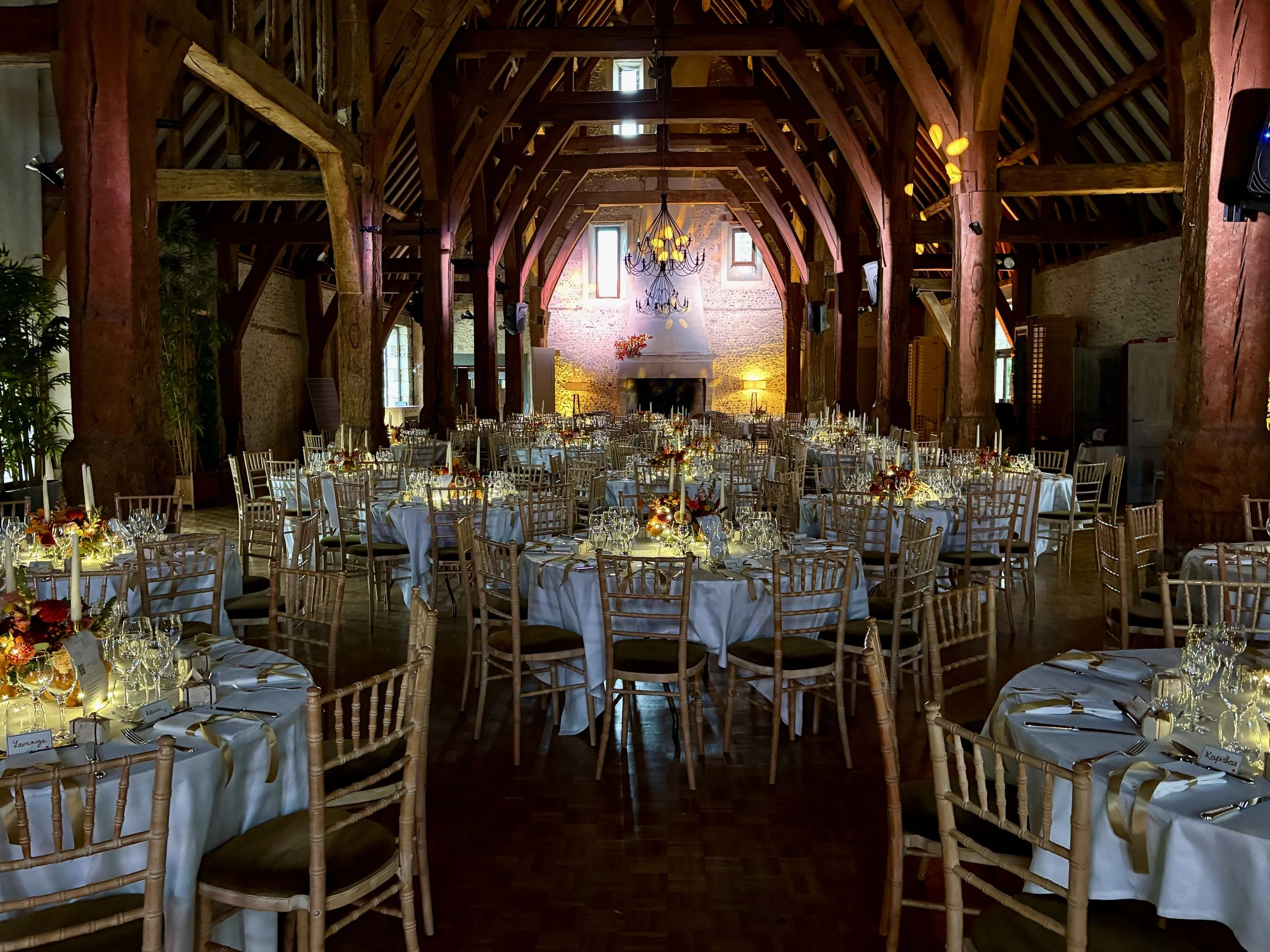Salle de réception décorée avec de grandes tables rondes, des chaises en bois, des chandeliers suspendus au plafond en bois, et des lampes d'ambiance, dans un bâtiment en pierre avec fenêtres naturelles.