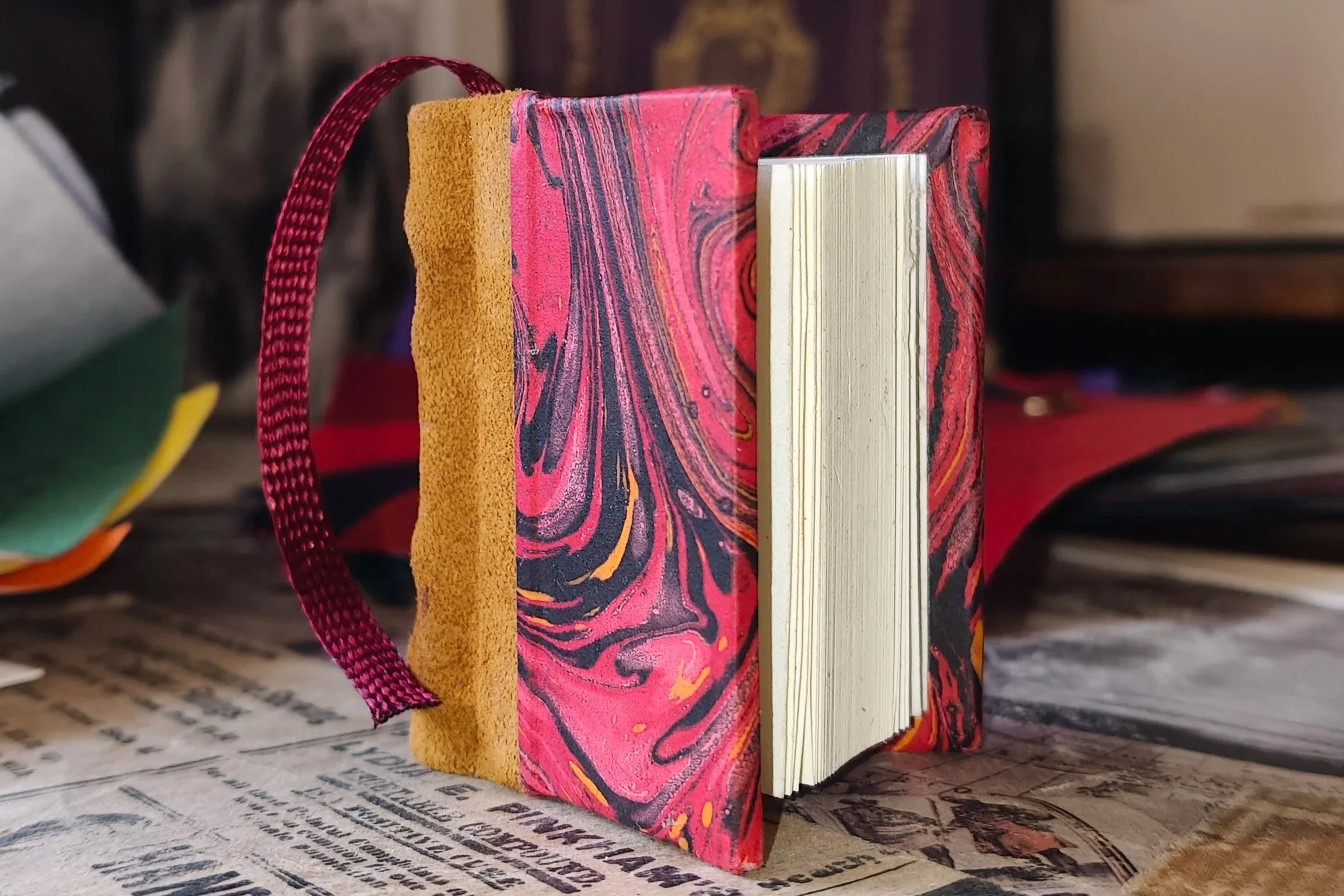 Mini libro rilegato con dorso in vera pelle, copertina in carta fiorentina rossa marmorizzata nei toni del nero e dell’oro, pagine dall’aspetto antico e segnalibro bordeaux.