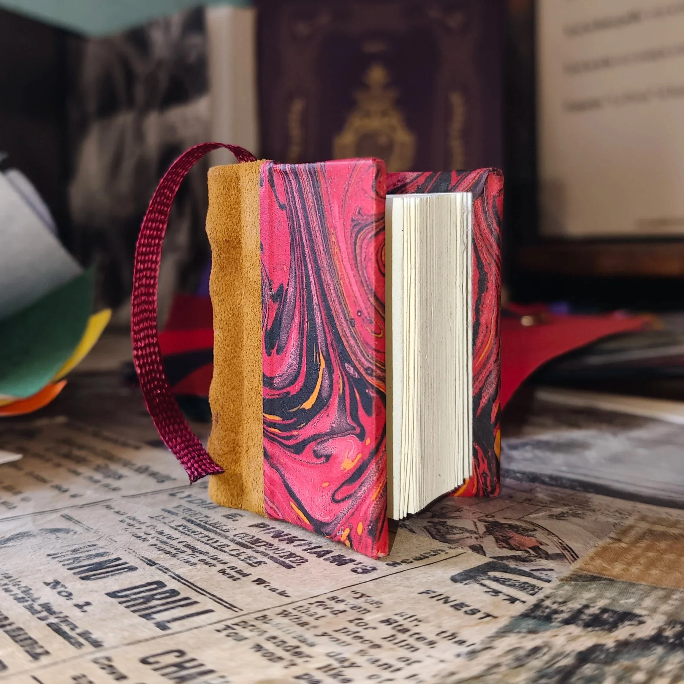 Mini libro rilegato con dorso in vera pelle, copertina in carta fiorentina rossa marmorizzata nei toni del nero e dell’oro, pagine dall’aspetto antico e segnalibro bordeaux.