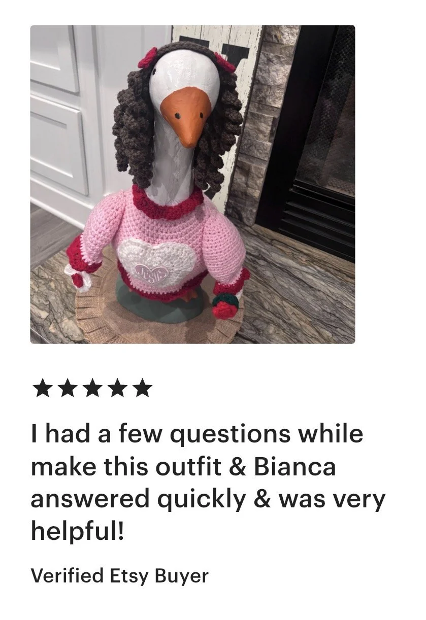 A decorative figure of a goose dressed in a pink knitted sweater with a heart design on the front, a red and white bracelet, and pink and red accessories on her head and neck. The figure is placed on a round mat on a kitchen countertop.