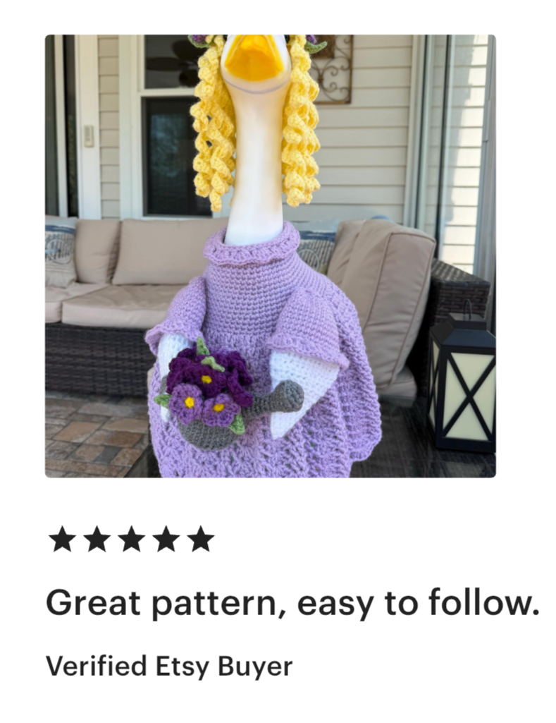 Handmade crochet decoration of a swan wearing a purple dress, with yellow yarn hair, holding a bouquet of purple flowers with yellow centers, placed outside against a porch with beige cushions and a black lantern.
