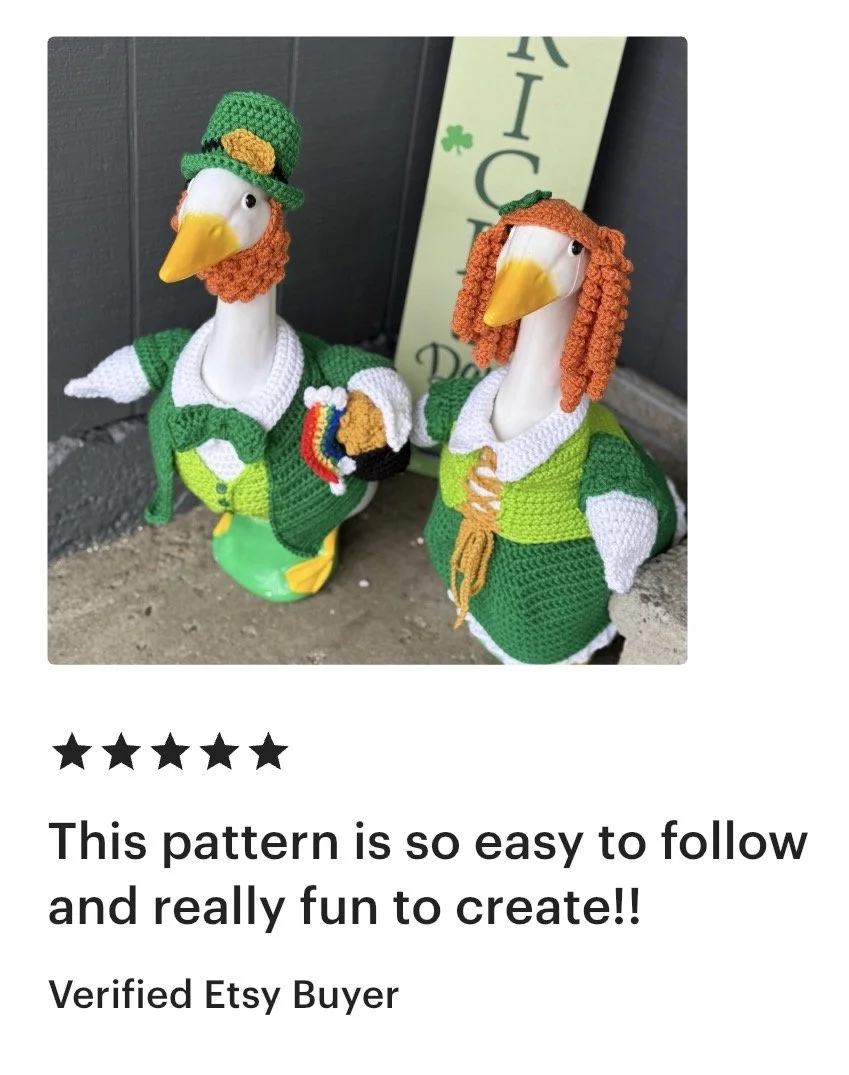Two geese figurines dressed in Irish-themed knit clothing, with one wearing a green hat and the other with curly orange hair, standing outdoors against a gray wall.
