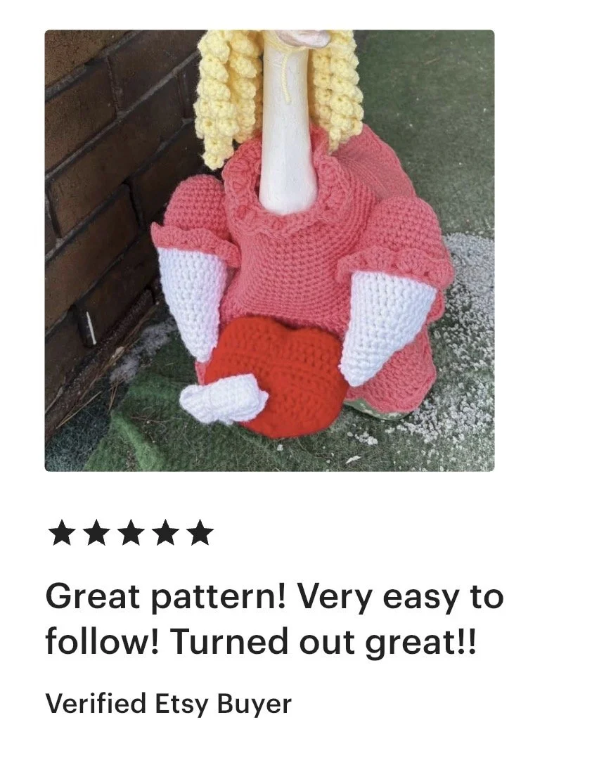 A crocheted figure resembling the Nutcracker character, with blonde hair, a pink coat, white gloves, and holding a red object, standing outdoors next to a brick wall.