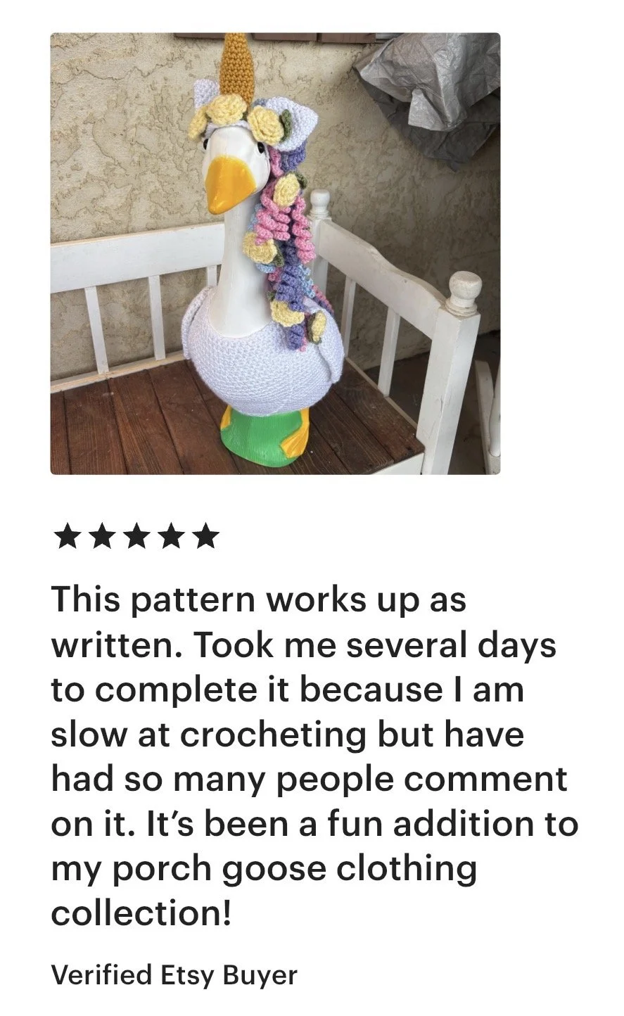 A porch goose sculpture decorated as a unicorn with a crocheted rainbow mane and horn, wearing a white crocheted garment, standing on a green base.