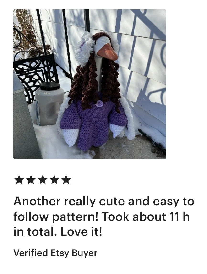 A knitted doll dressed as a stork, with brown curly yarn hair, wearing a purple dress and white gloves, sits outside on snow next to a black metal chair, a white container, and a white wall with shadows.