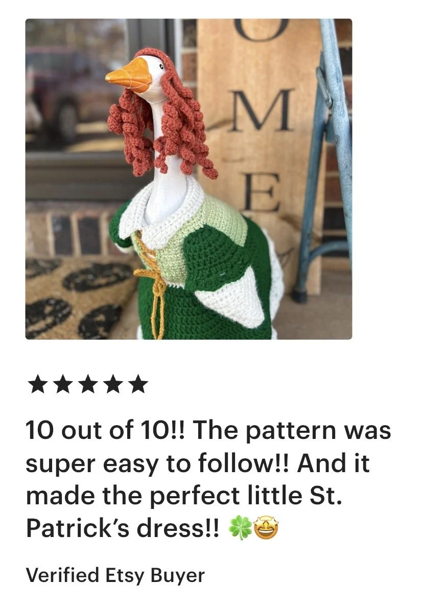 A knitted and crocheted stork wearing a green and white St. Patrick's Day dress with a white collar and yellow ribbon, displayed outdoors beside a wooden sign with black lettering.