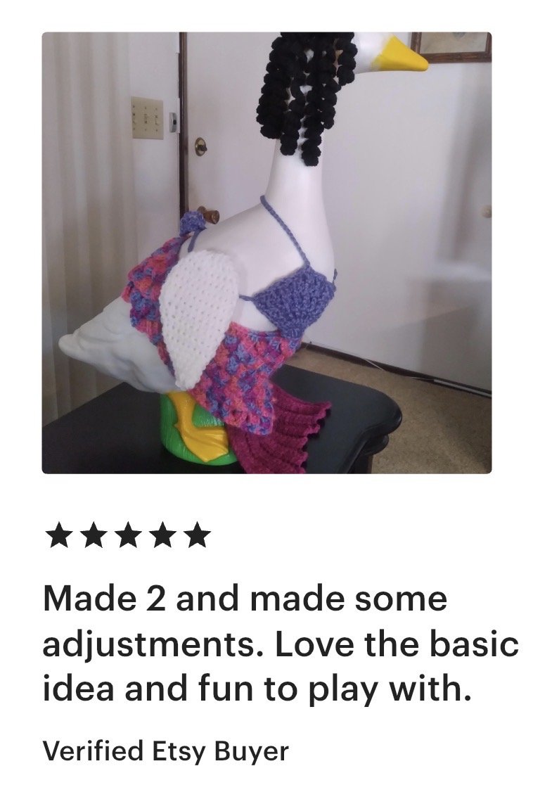 A decorative sculpture of a duck dressed with handmade knitted accessories, including a purple bikini top, a multicolored skirt, and a maroon skirt. The duck has a yellow beak, a black curly hair wig, and is standing indoors on a table.