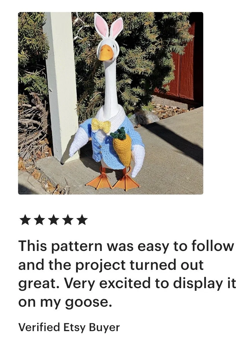 A decorative goose statue dressed as a spring or Easter character with bunny ears, a yellow bow tie, and a blue jacket, holding a felt carrot, standing outside in front of green shrubbery and a red wooden fence.