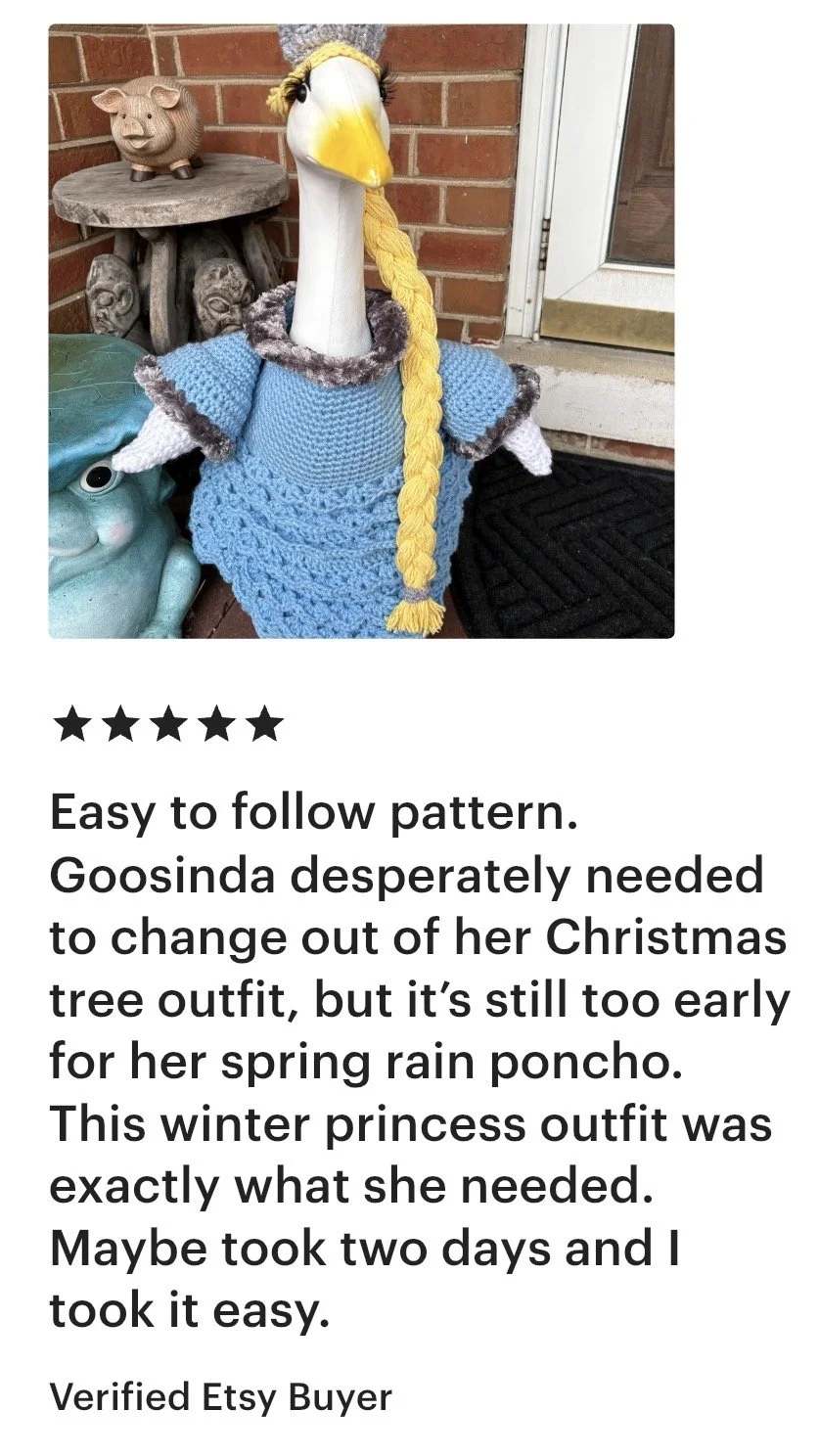 A stuffed goose dressed in a blue knit winter outfit with gray trim and a yellow braid, standing outdoors on a brick porch next to a pig-shaped garden ornament and a ceramic frog statue.