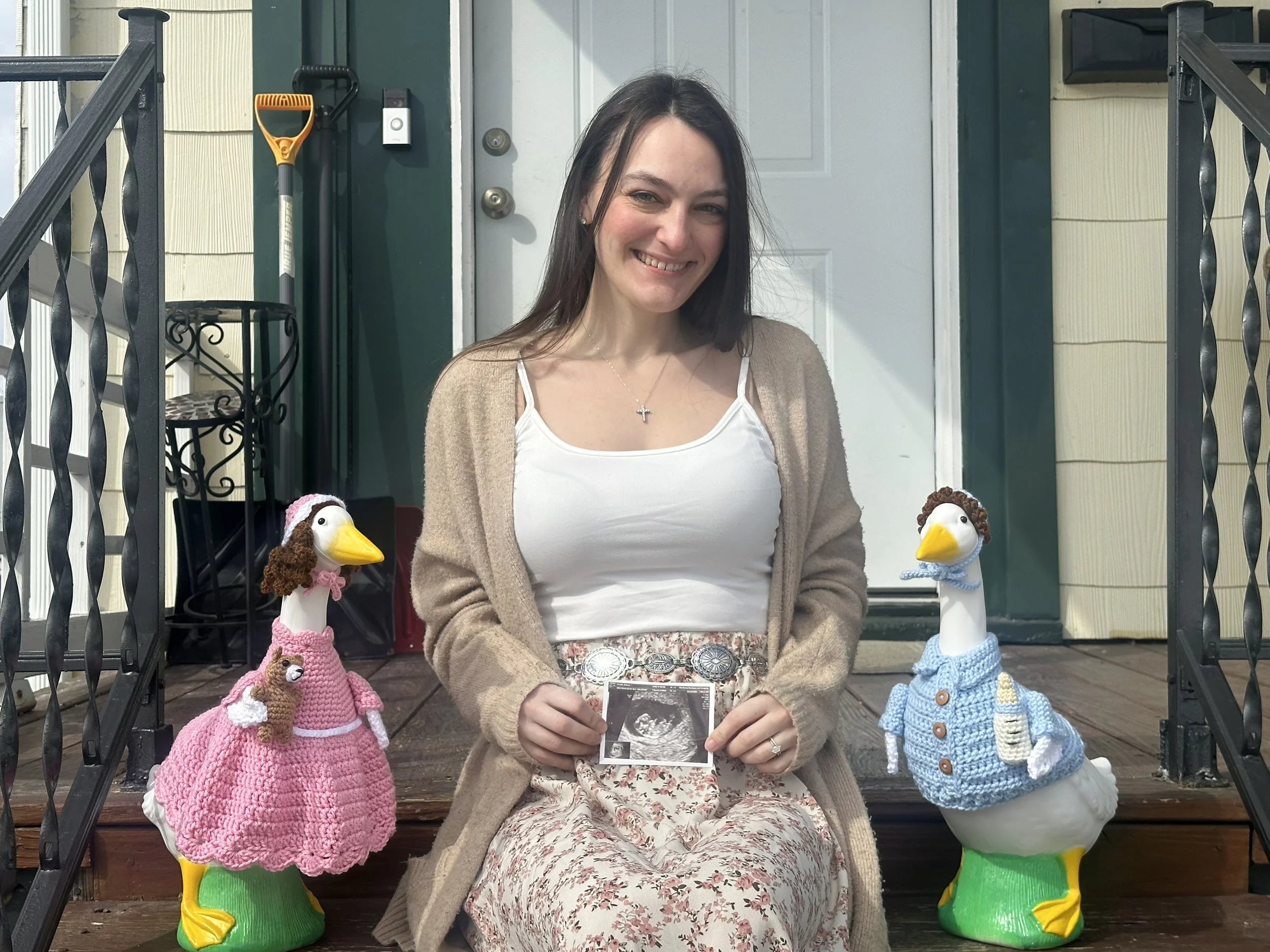 A woman sitting on a porch holding an ultrasound picture, with two duck-decorated chairs beside her, and gardening tools and a door in the background.