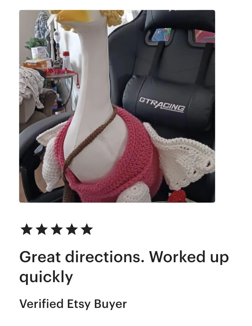 A white plastic giraffe figurine wearing a pink and white knitted dress with a brown strap, placed on a black ergonomic chair in a room with cluttered furniture and a gray GTRACING gaming chair in the background.