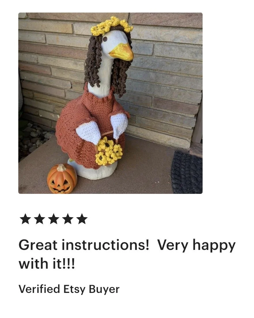 Kinetic sculpture of a goose dressed in crochet clothing with a flower crown, holding a bouquet, with a pumpkin-shaped jack-o'-lantern decoration nearby.