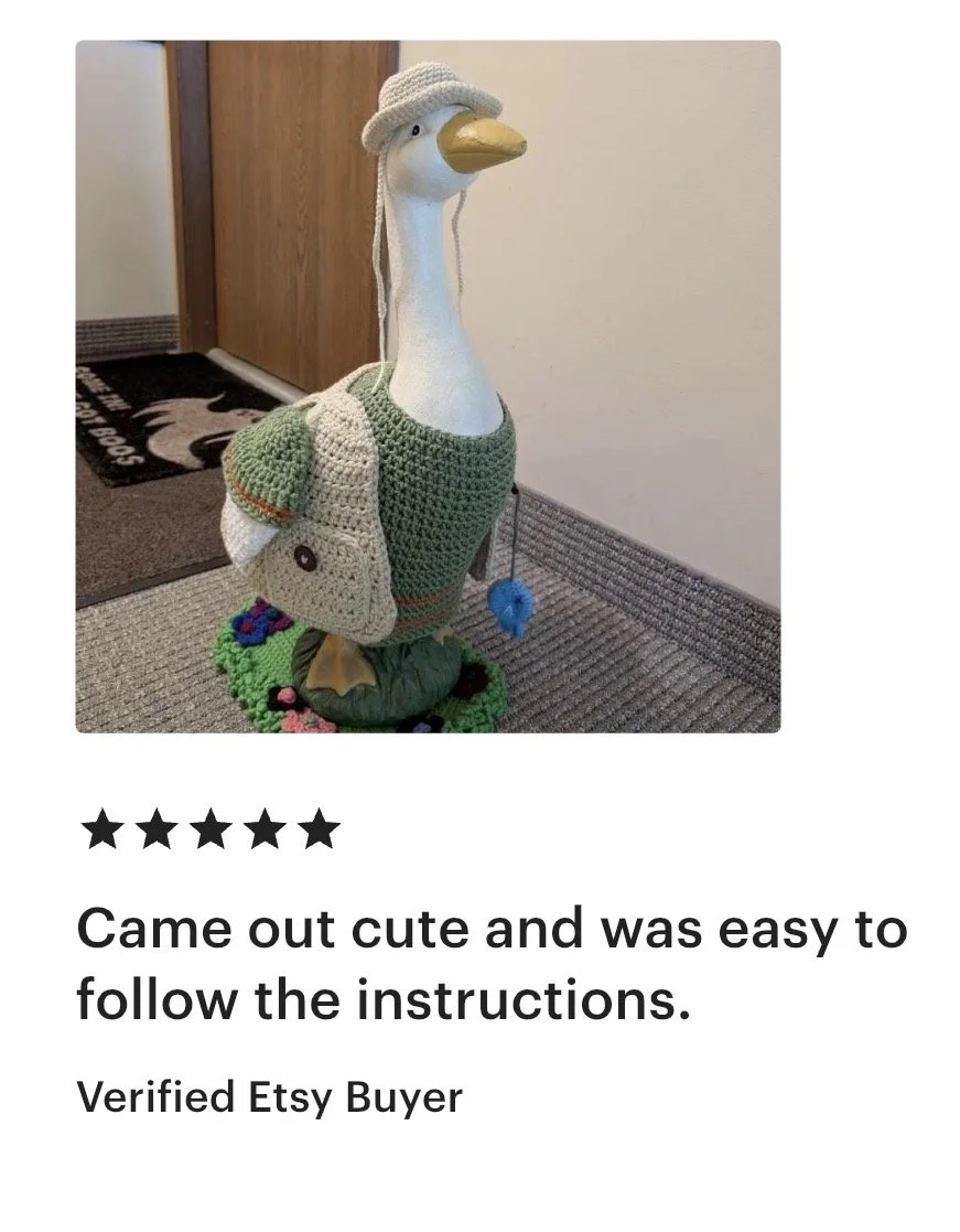 A decorative goose statue dressed in a crochet outfit, including a hat, dress, and a bag, standing on a doll with grass and flower decorations on the floor.
