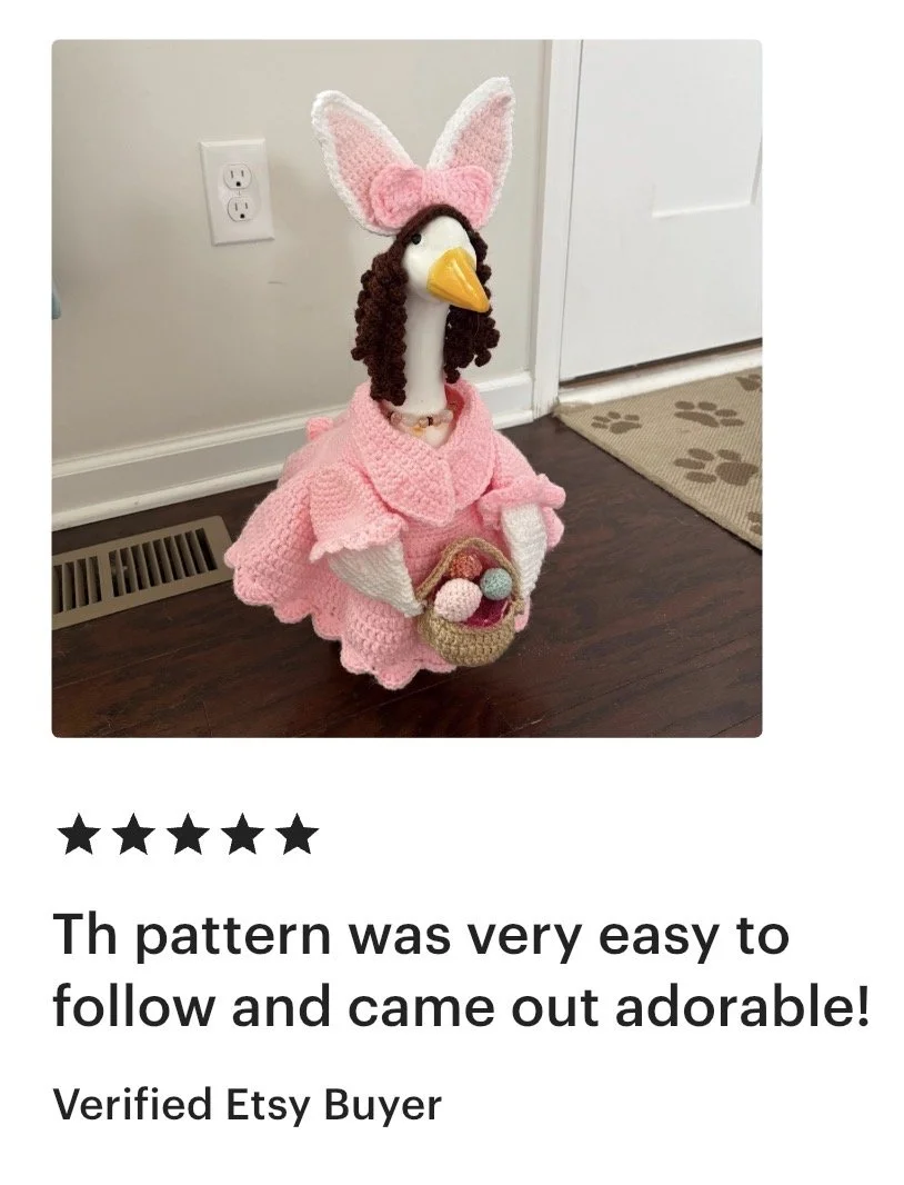 A handcrafted stuffed animal resembling a goose dressed in pink crocheted clothing, with bunny ears on its head and holding a small basket filled with colorful pom-poms.