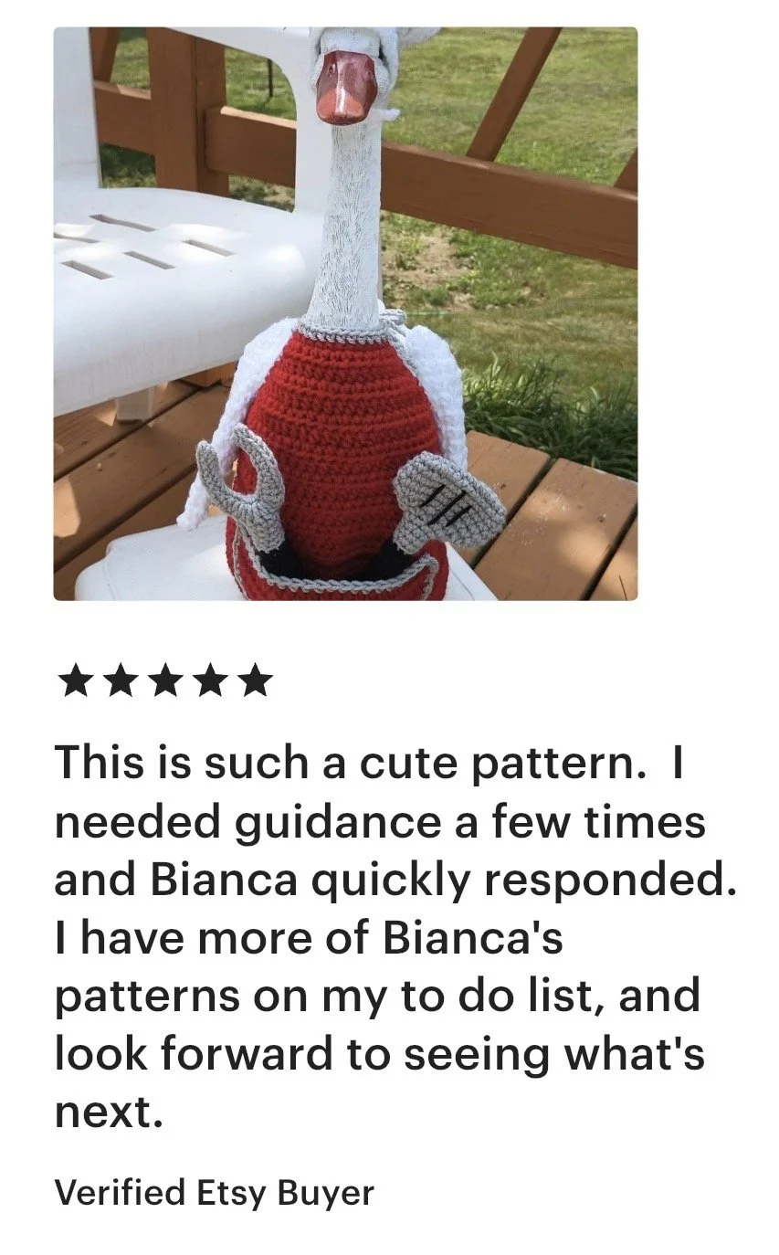 A crocheted or knitted plush toy penguin with a white face and orange beak, wearing a red sweater, sitting outdoors on a wooden bench with a white plastic chair nearby and green grass in the background.