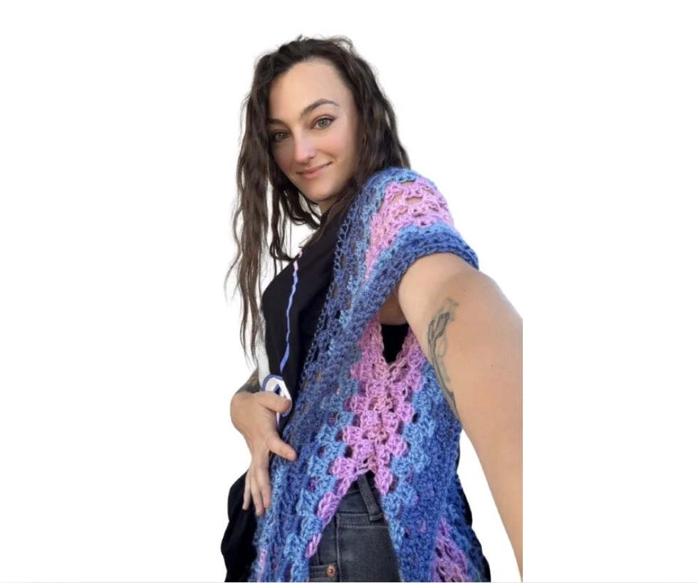 A woman with long, wavy brown hair and light skin taking a selfie. She is wearing a colorful crocheted shawl in shades of pink, purple, and blue, along with a black top and jeans. She has a tattoo on her upper arm and is smiling at the camera against a plain white background.