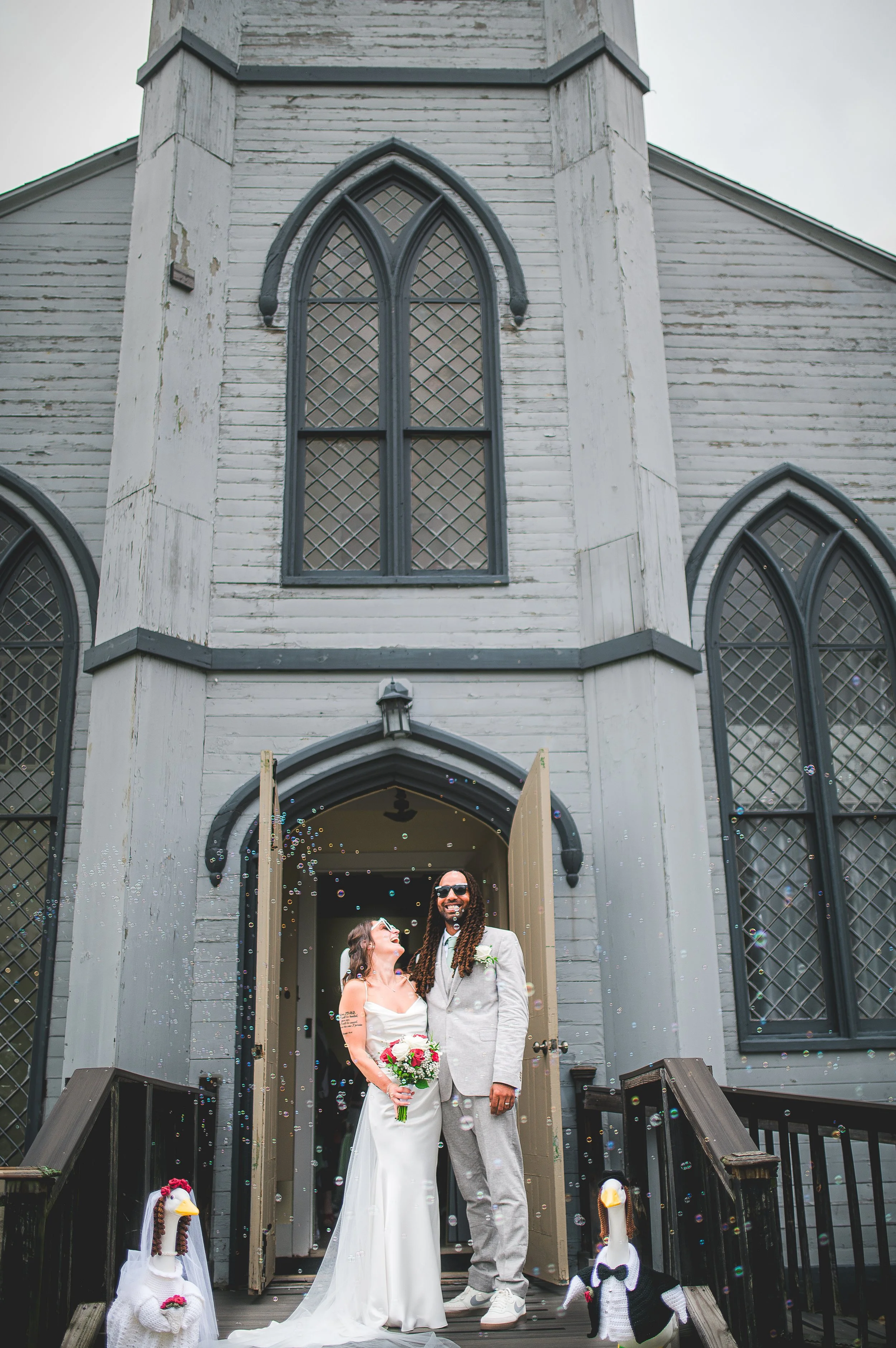 A wedding couple stands in front of a church entrance, smiling and looking at each other, with two large decorative duck figures dressed as a bride and groom in front of them. The bride wears a white wedding gown and holds a bouquet, while the groom wears a light gray suit.