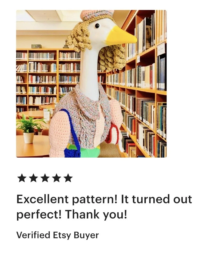A large puppet resembling a goose dressed in a crocheted outfit standing in a library with bookshelves filled with books in the background.