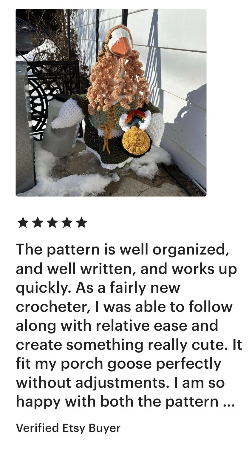 A crocheted porch goose holding an apple, standing on snow near a white wall, with a metal table and a propane tank nearby.
