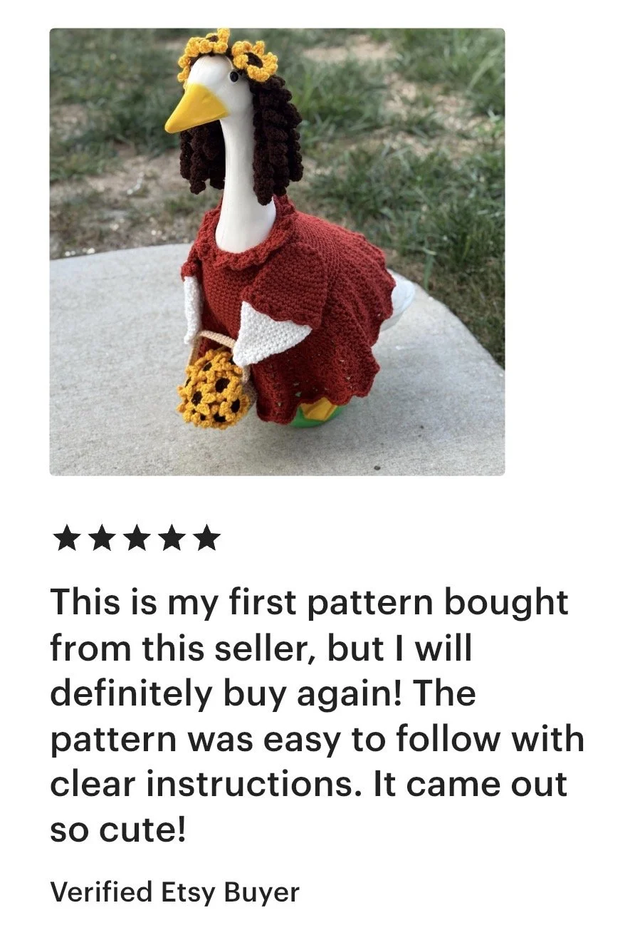 A goose-shaped toy dressed in red knitted clothing with a brown and yellow floral hat, holding a small bouquet of knitted yellow flowers, placed outdoors on a concrete surface.