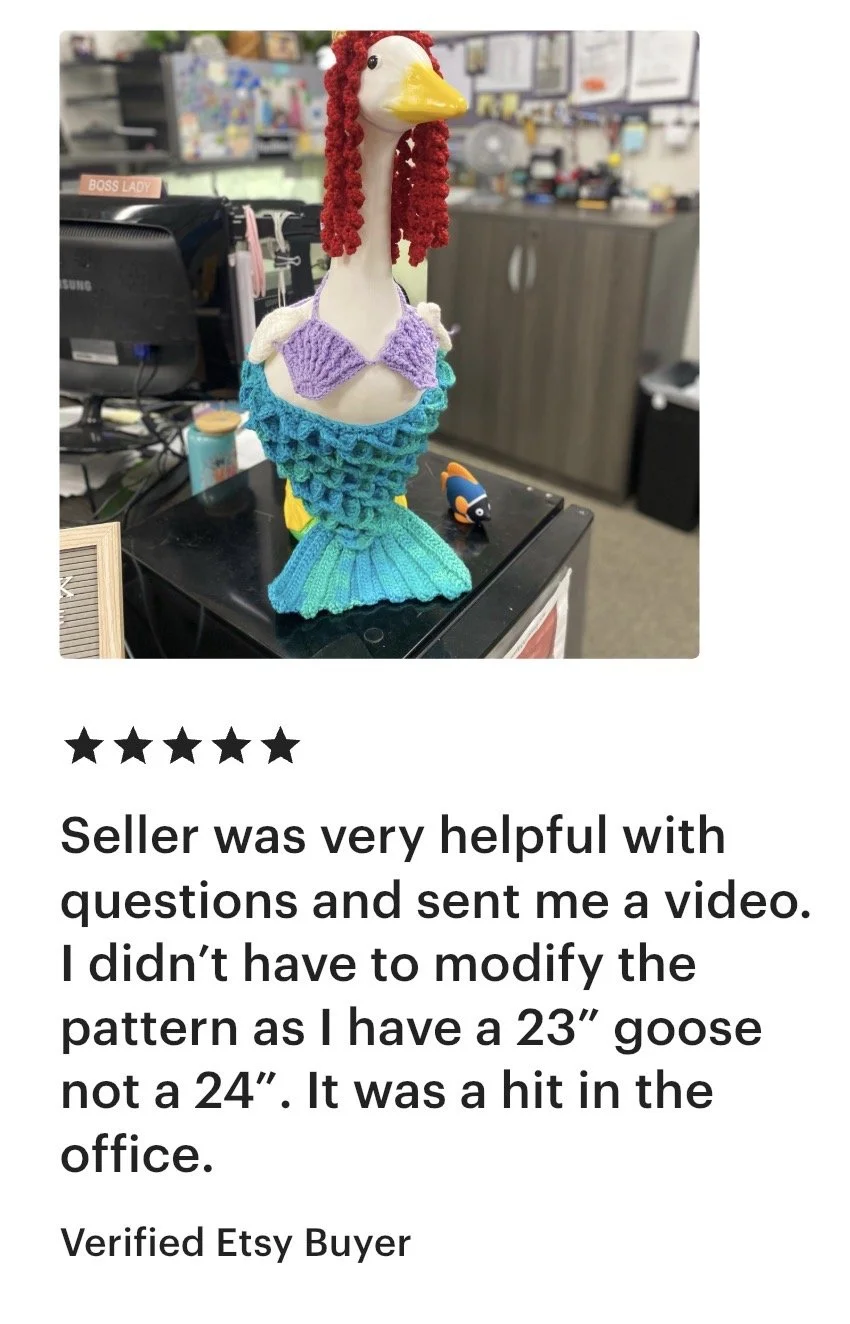 A handmade knitted goose with a white body, red curly hair, yellow beak, purple bikini top, and turquoise tail, placed on a black surface in an office setting.