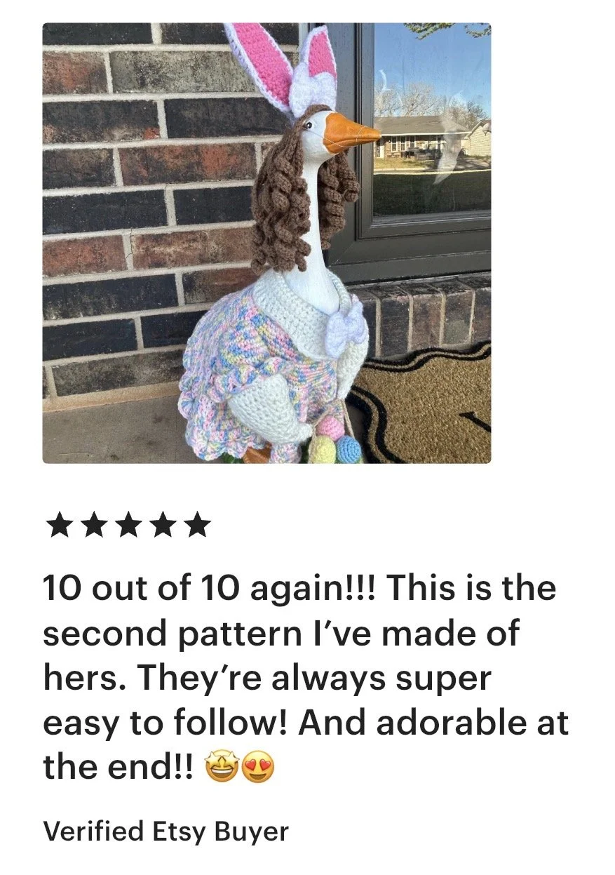 A stuffed fabric doll of a goose dressed in a colorful, knitted outfit with bunny ears hat, standing outside near a brick wall and a glass door.