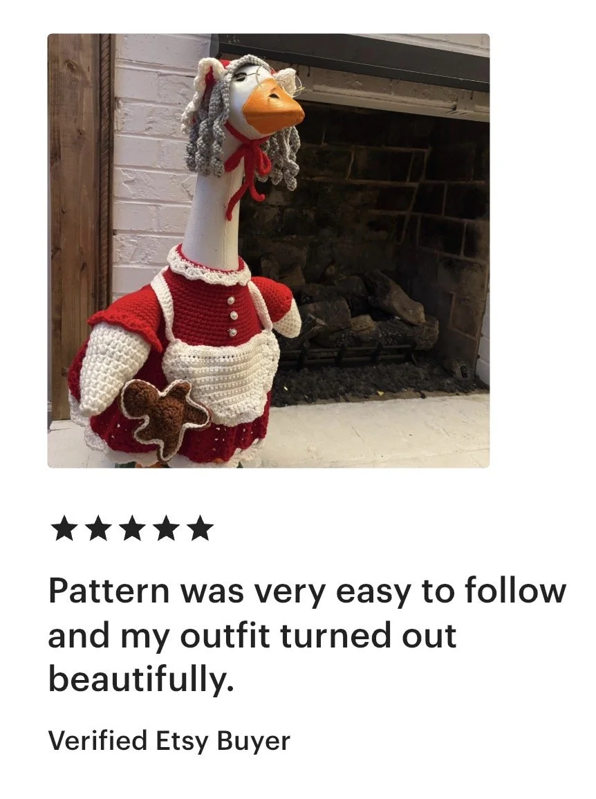 A life-sized stuffed goose dressed in a red and white crocheted Christmas outfit, holding a gingerbread man, standing near a fireplace.