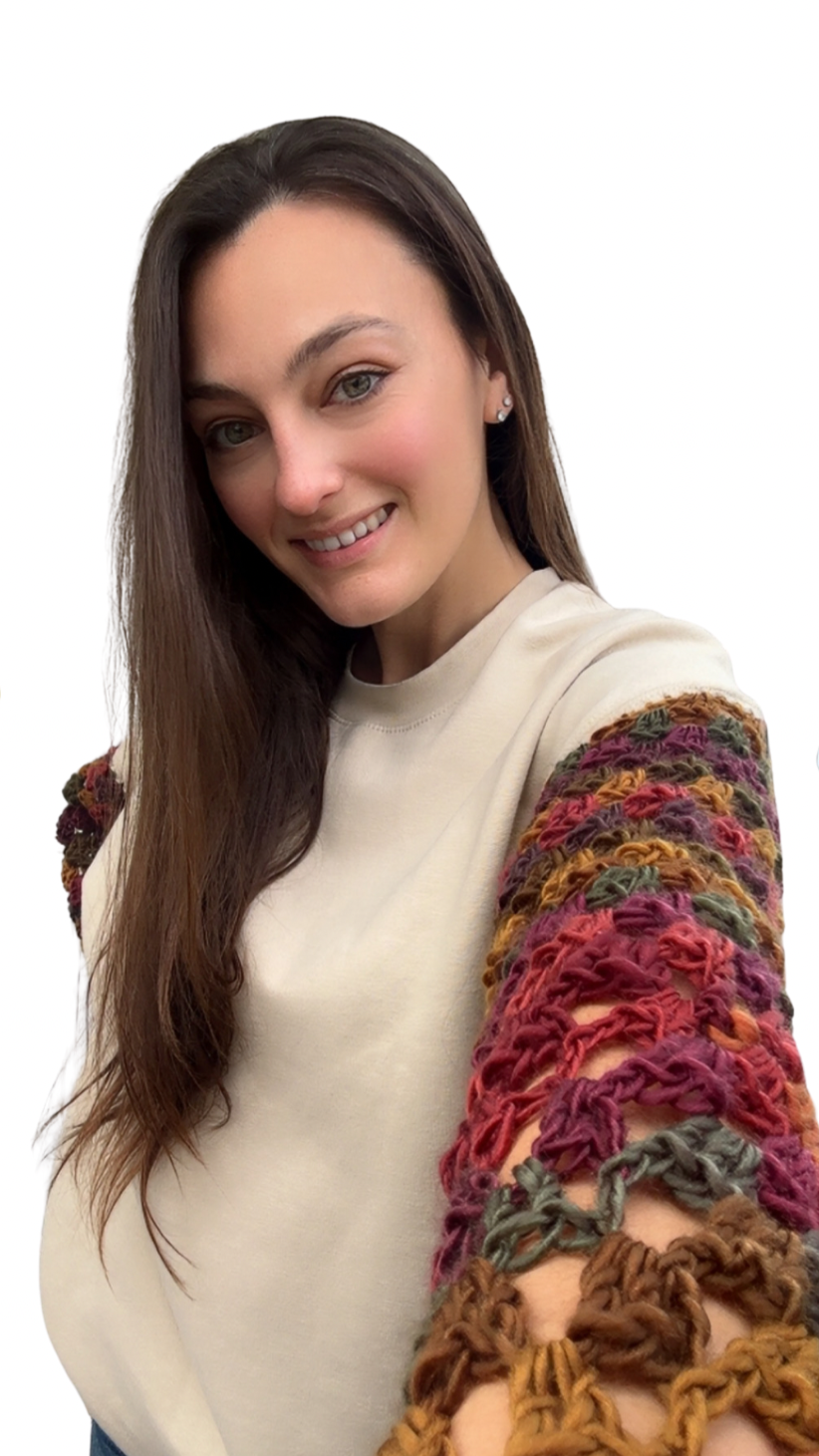 A young woman with long brown hair, green eyes, and a friendly smile taking a selfie against a plain white background. She is wearing a cream-colored sweatshirt with colorful, textured, open-knit sleeves in shades of red, pink, green, and brown.