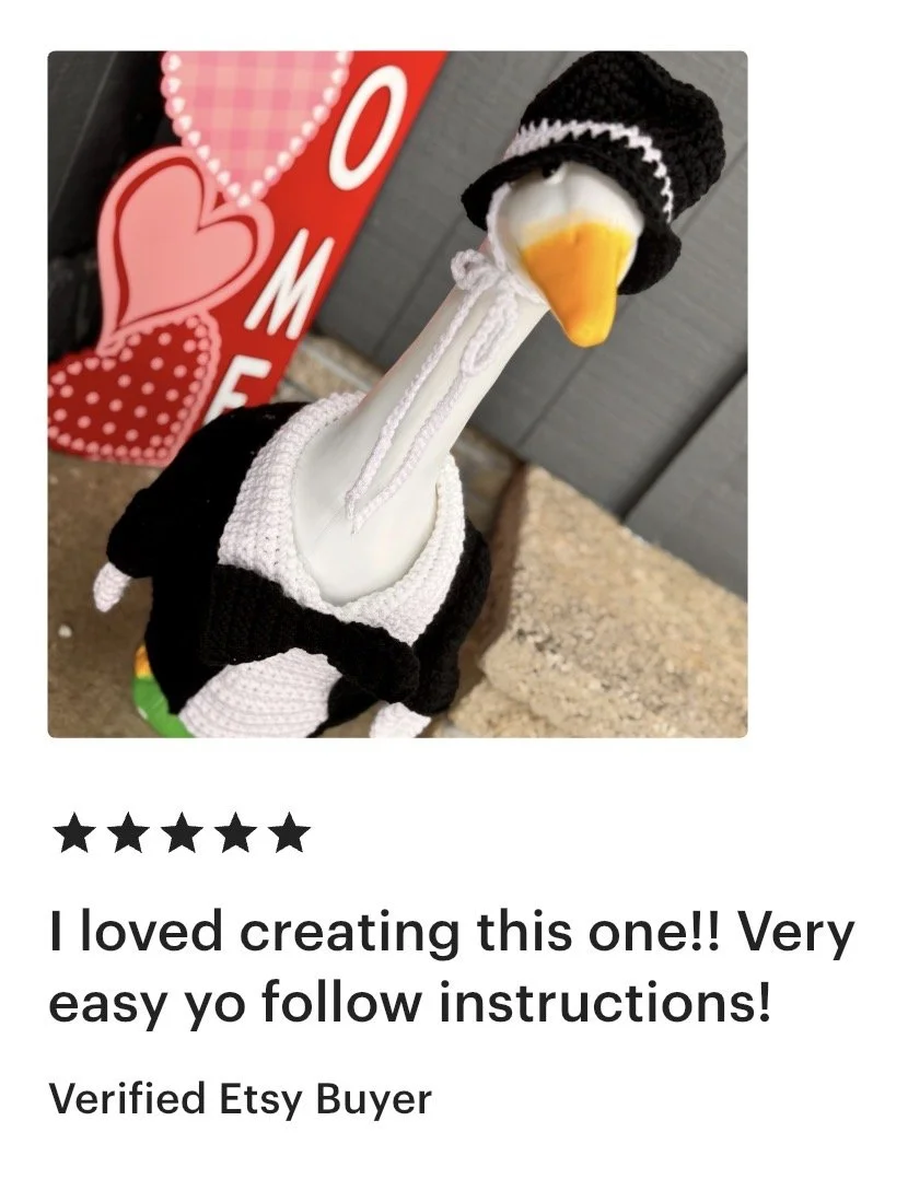 A decorative figure of a goose dressed in a black sweater and a black knit hat with a white trim. The goose has a yellow beak and is wearing a white scarf. Behind it, there is a red card with the word "LOVE" and hearts, indicating a Valentine's Day o